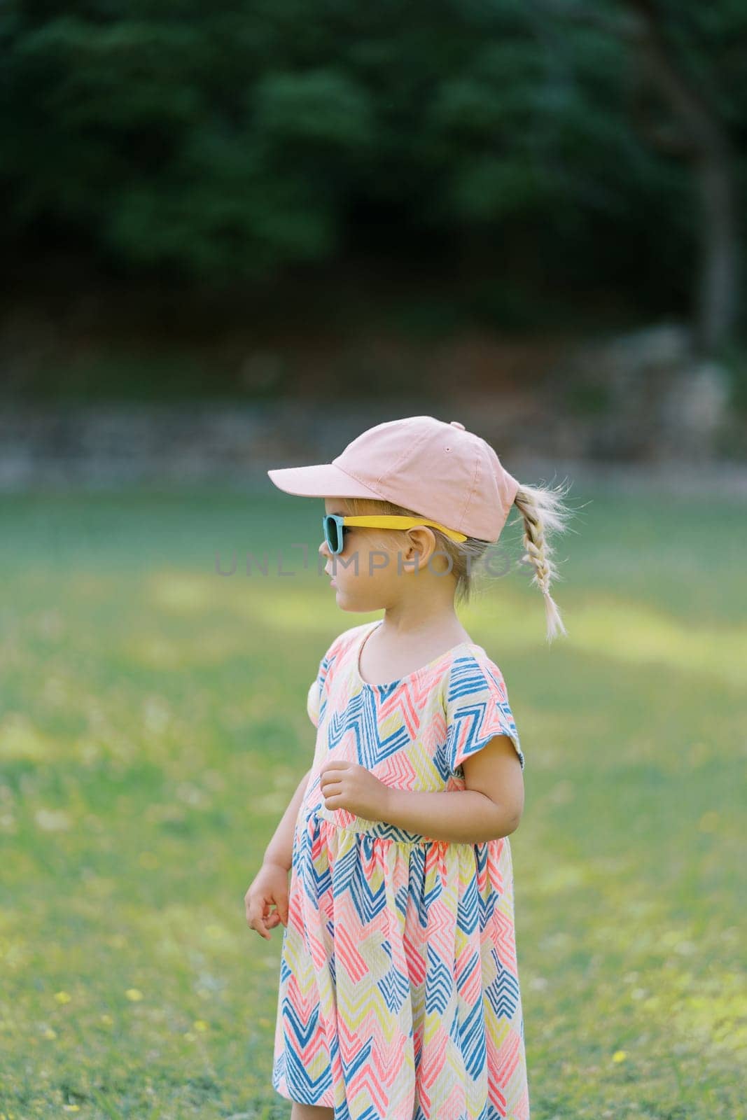 Little girl in sunglasses stands on the lawn looking into the distance. Side view. High quality photo