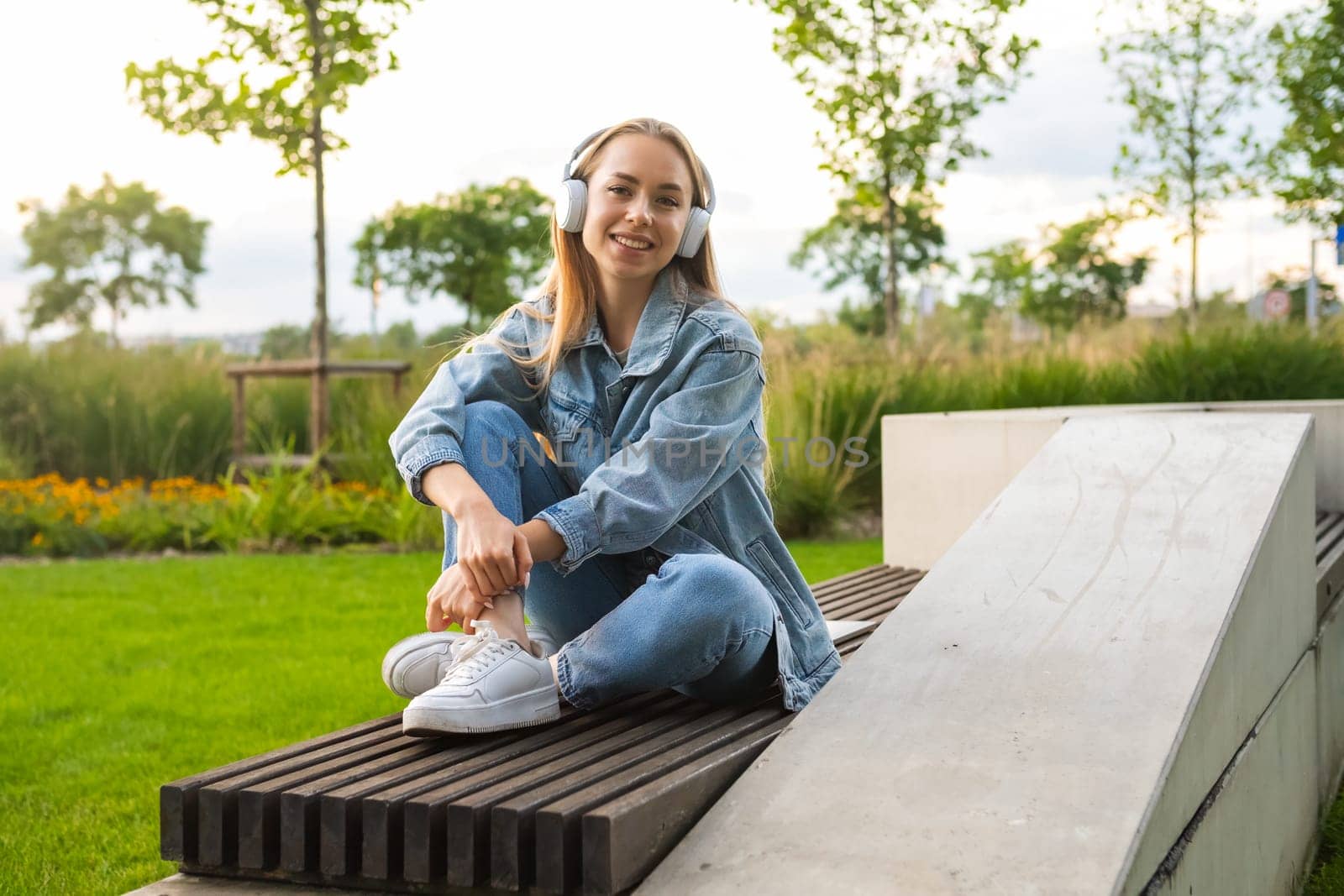 Fair haired girl is seated on a park bench with legs crossed and listening to music via headphones by vladimka