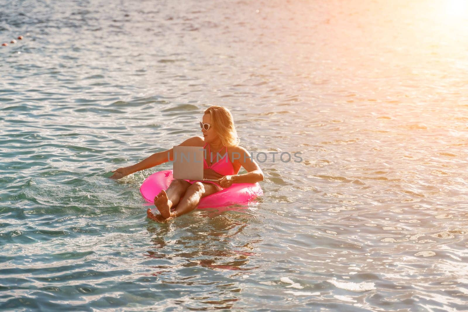 Woman works on laptop in sea. Freelancer, blond woman in sunglases floating on an inflatable big pink donut with a laptop in the sea at sunset. People summer vacation rest lifestyle concept