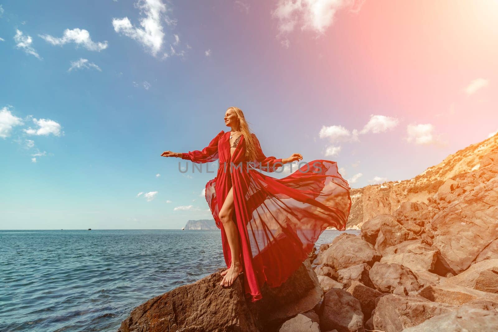 Red dress sea woman. A blonde with flowing hair in a long flowing red dress stands on a rock near the sea. Travel concept, photo session at sea by Matiunina