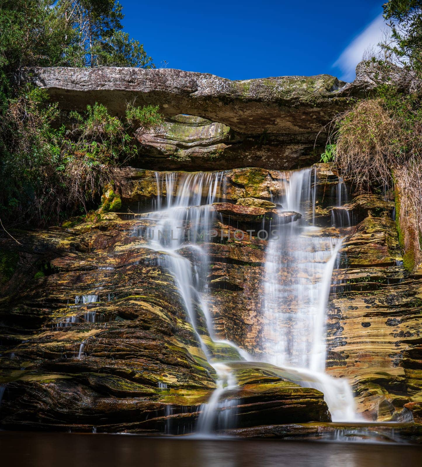 Beautiful nature scene featuring a silky waterfall cascading over a rock wall beneath an overhanging rock with a hole.