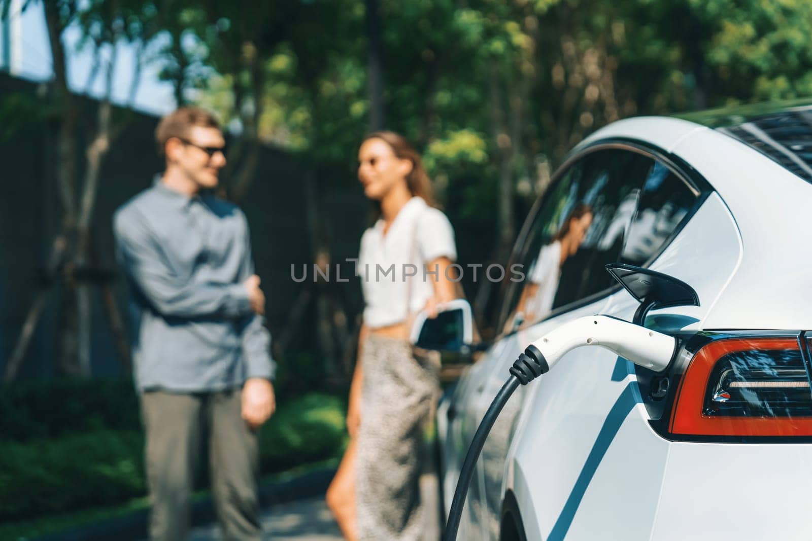 Young couple travel with EV electric car charging in green sustainable city outdoor garden in summer shows urban sustainability lifestyle by green clean rechargeable energy of electric vehicle innards