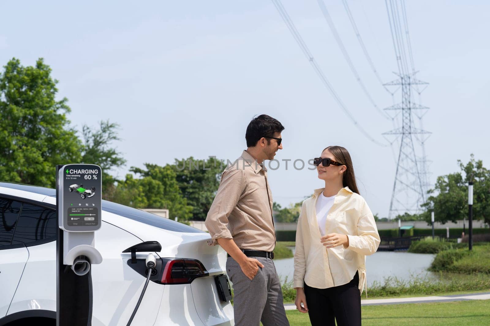 Young couple recharge EV car battery at charging station connected to power grid tower electrical industrial facility as electrical industry for eco friendly vehicle utilization. Expedient