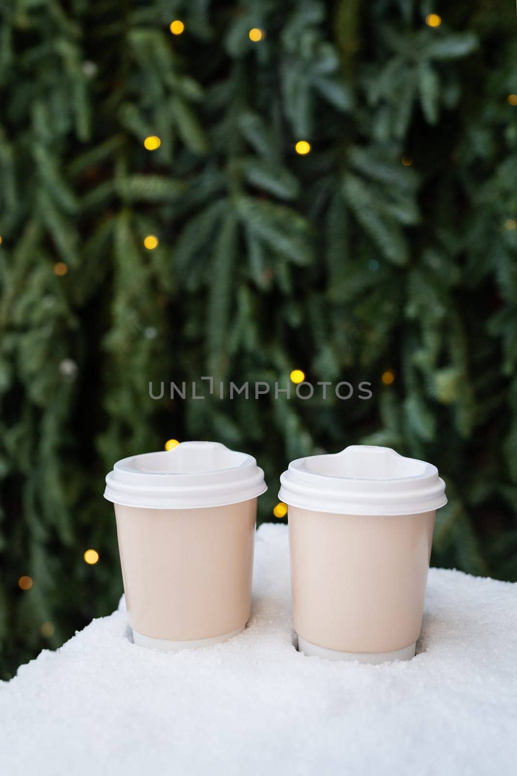 Two paper coffee cups on a snowy surface, with a Christmas tree with lights in the background
