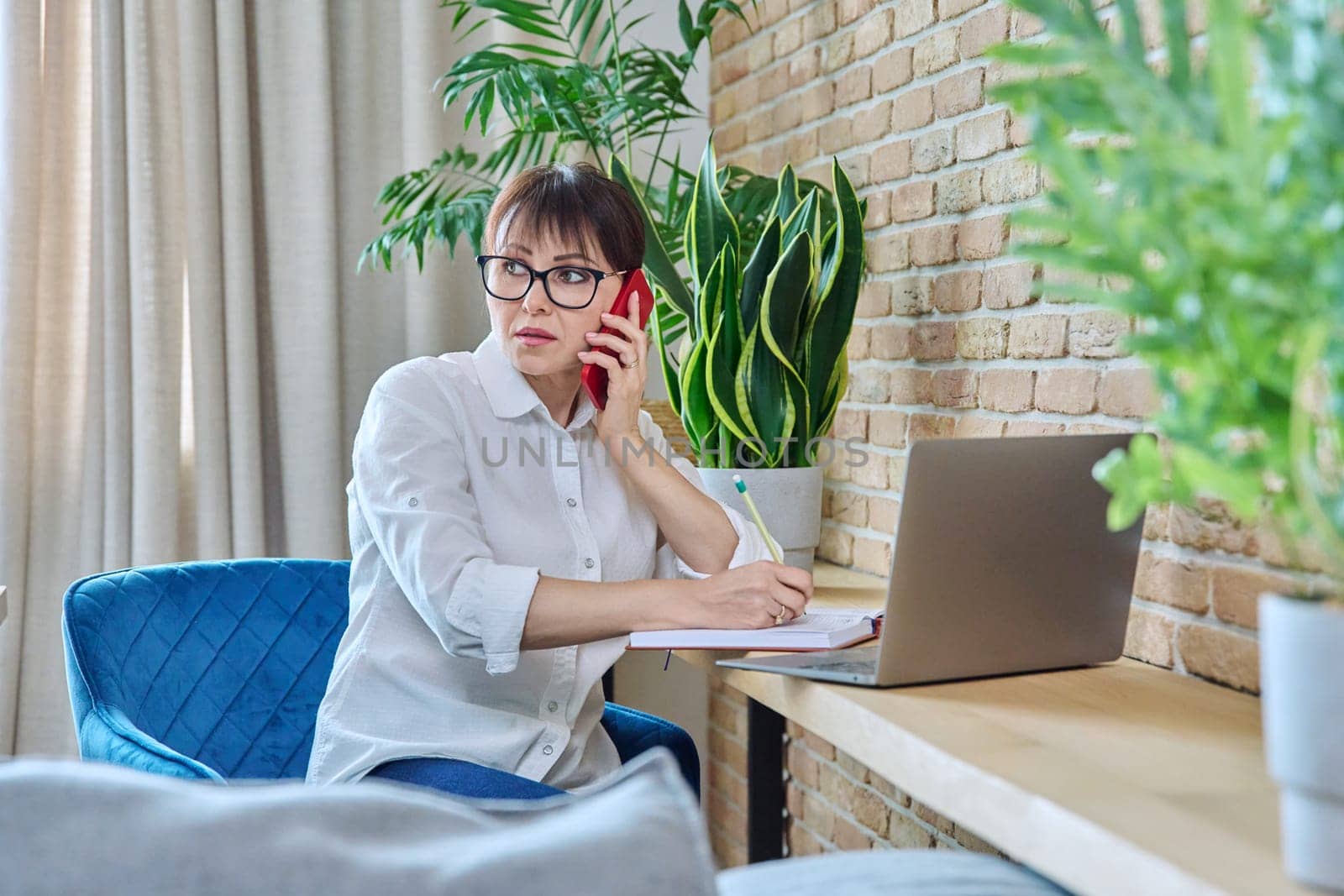 Middle-aged woman talking on mobile phone, sitting at desk at home with laptop computer. Female working remotely, freelancing, online services, technologies for work communication leisure