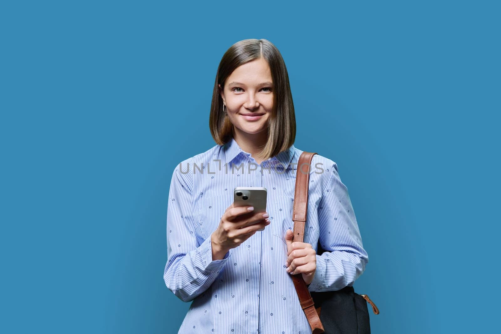 Young woman student with smartphone backpack on blue studio background. Smiling attractive female looking at camera. Using mobile applications apps for work business study leisure, technology, people