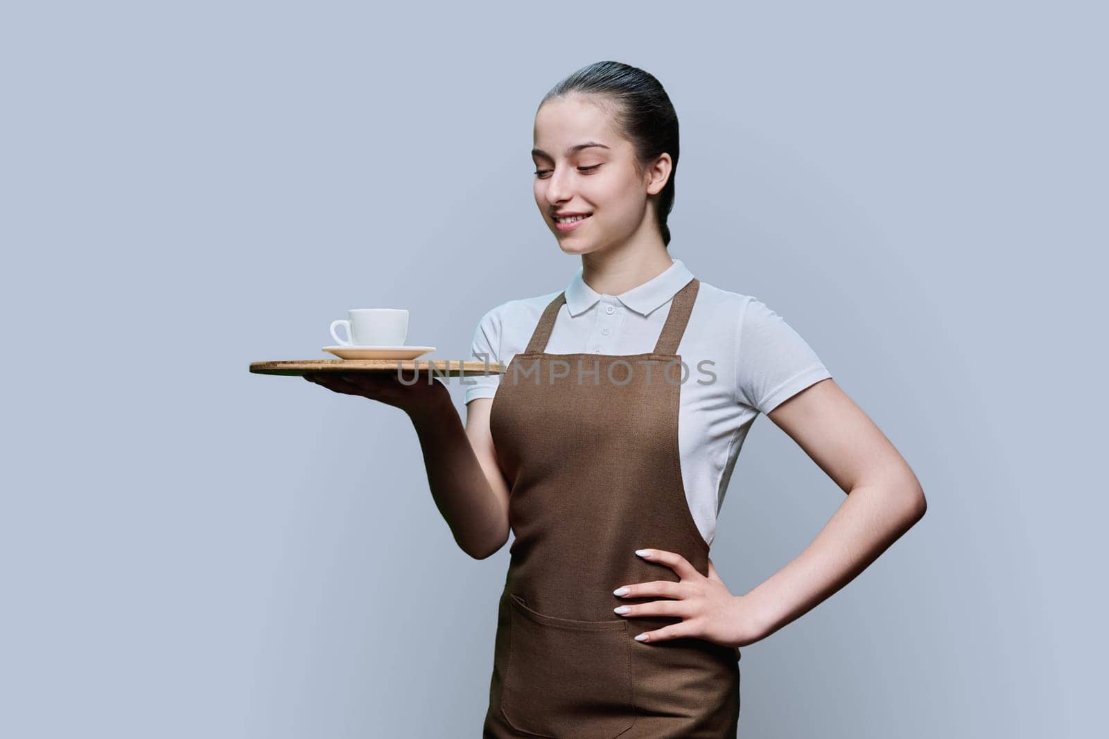 Portrait of young female waitress in an apron with tray of cup of coffee on gray studio background. Smiling girl looking at tray with order. Work, service in food, youth concept