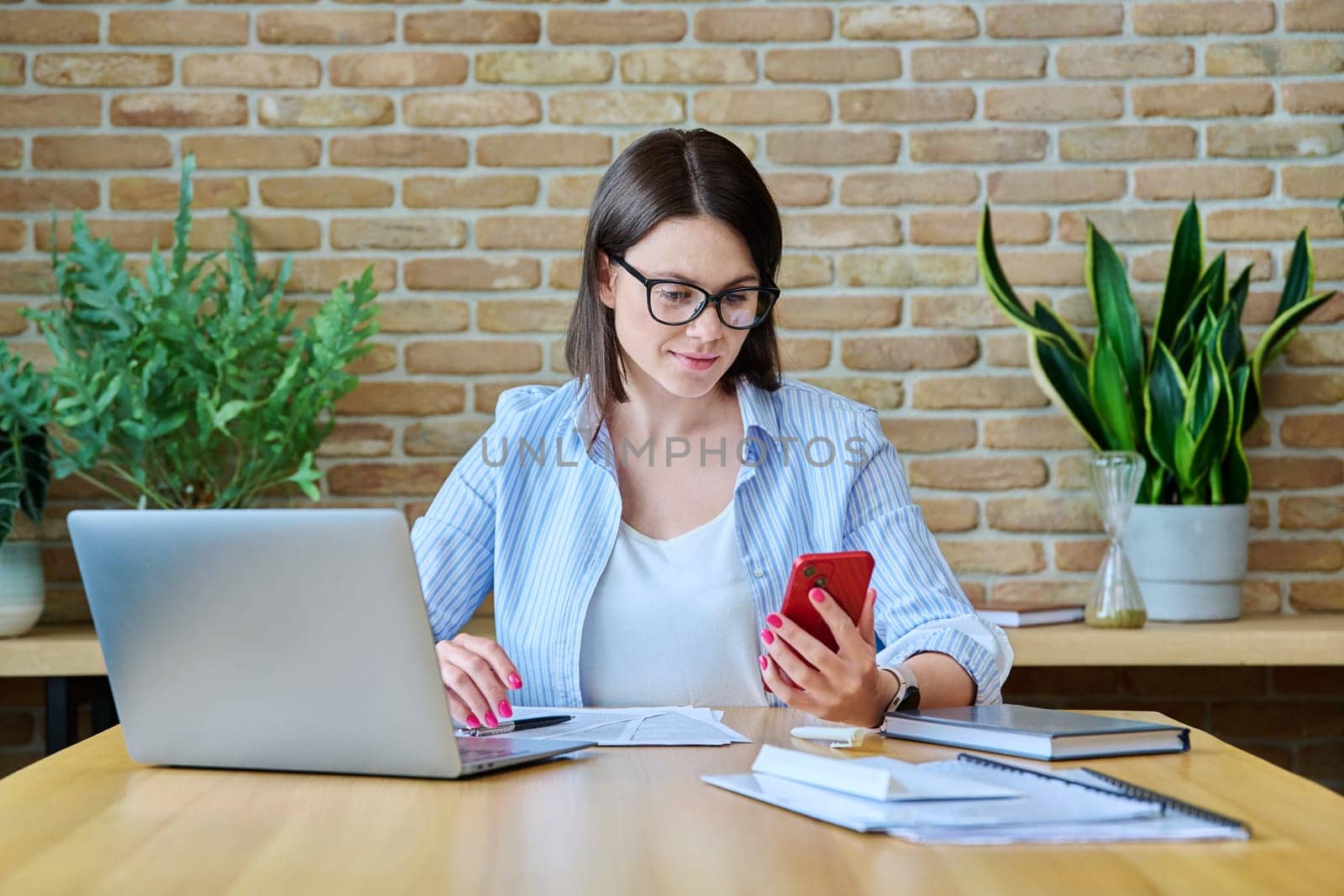 Young business woman sitting at desk with computer in office, using smartphone by VH-studio