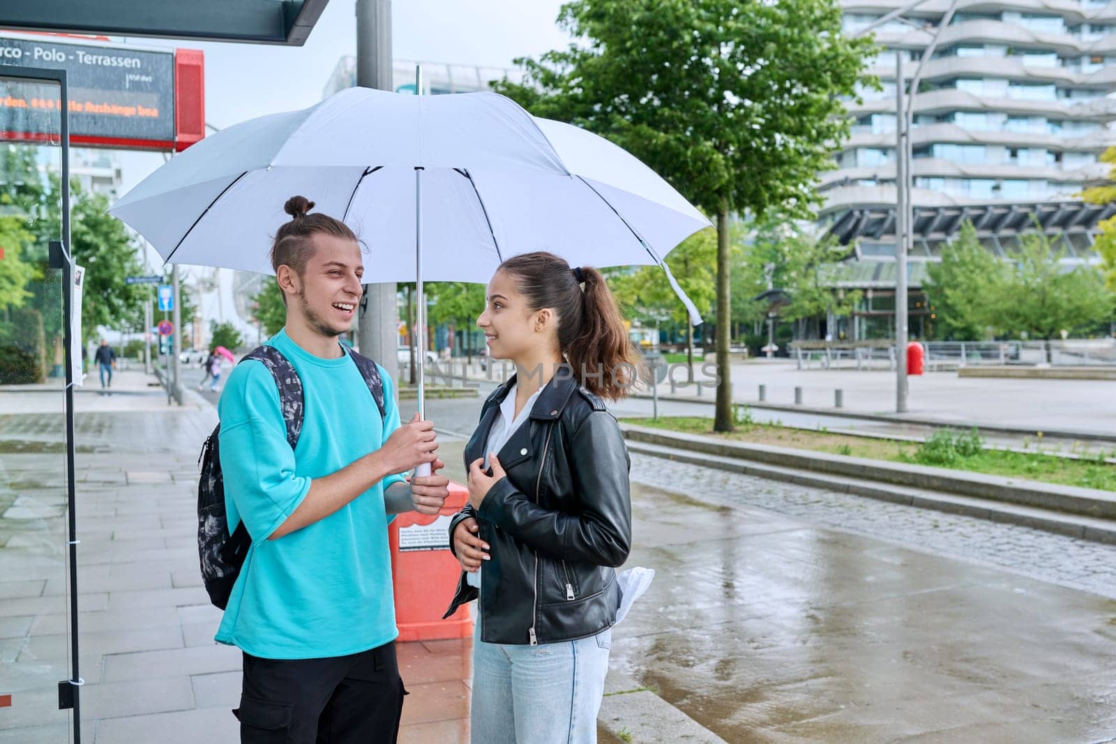 Teenagers guy and girl students with backpacks at bus stop under umbrella waiting for city bus, road copy space, in rainy weather. Public transport, adolescence youth, city life lifestyle, urban style