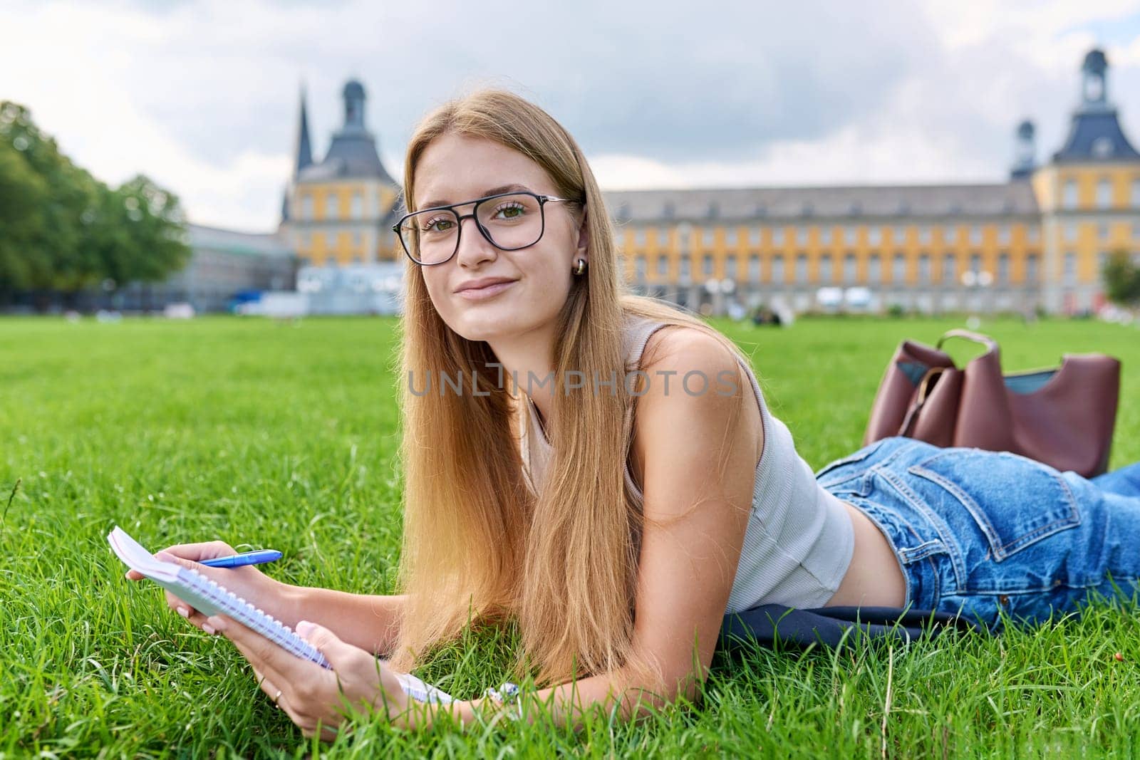 Portrait of young teenage girl student lying on lawn on grass, with textbook notebook, looking at camera, educational building background. Youth, education, college university concept