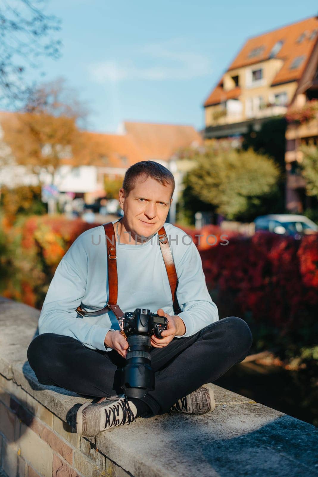 Man Sitting in Old European City And Holding Photo Camera. Contemporary Stylish Blogger And Photographer. Handsome man taking a selfie on a trip in Europe. by Andrii_Ko