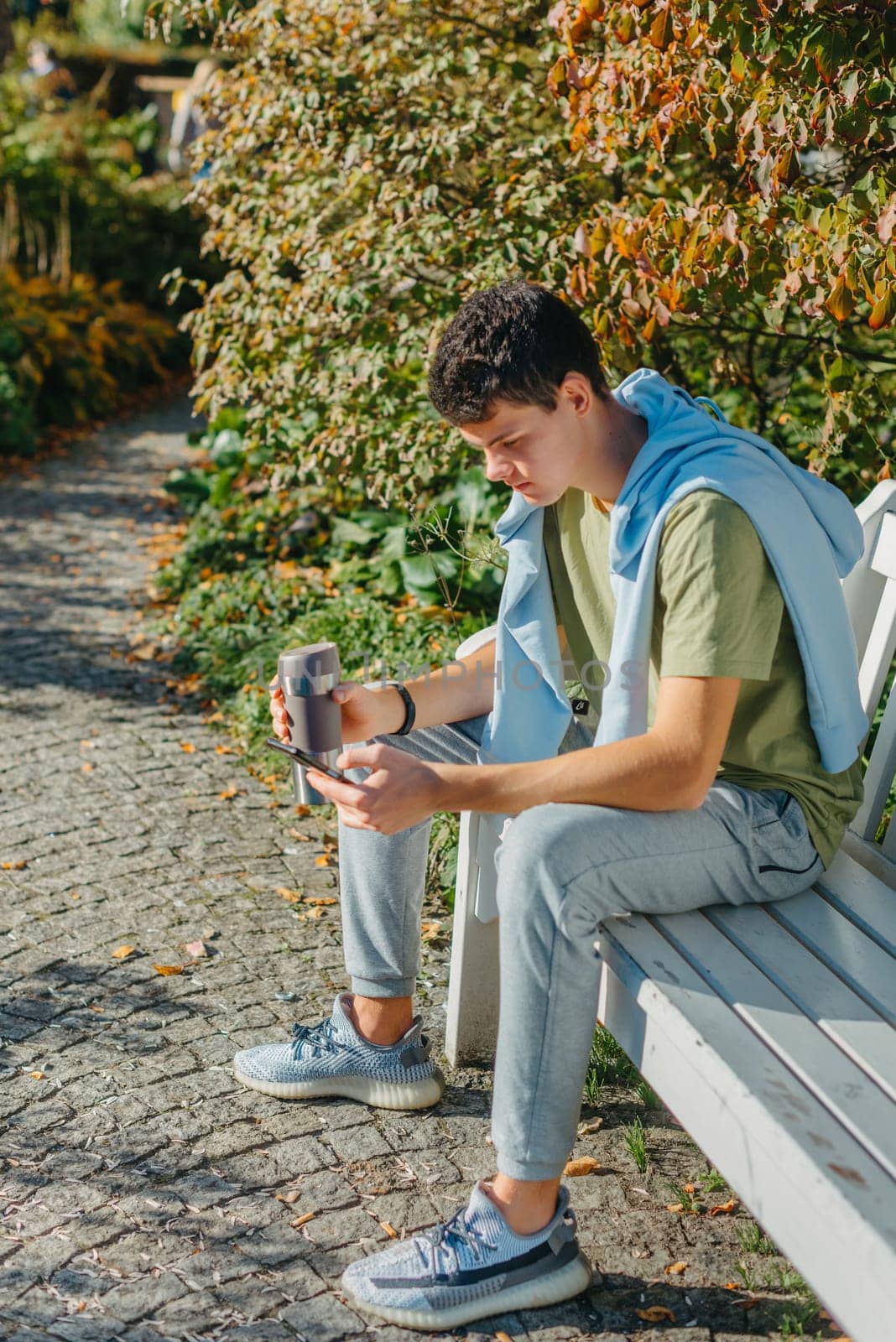 A Teenager Sits On A Bench In The Autumn Park Drinks Coffee From A Thermo Mug And Looks Into A Phone. Portrait Of Handsome Cheerful Guy Sitting On Bench Fresh Air Using Device Browsing Media Smm Drinking Latte Urban Outside Outdoor by Andrii_Ko