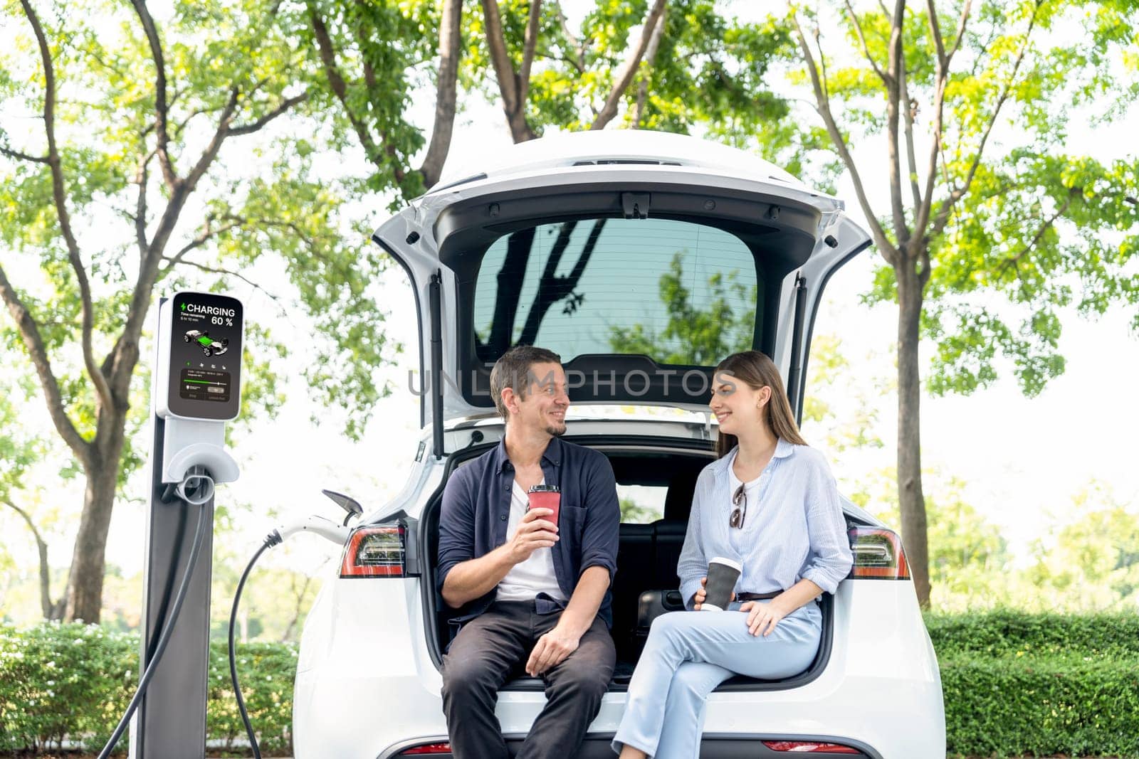 Lovely young couple drinking coffee while recharging battery for electric car during road trip travel EV car in natural forest or national park. Eco friendly travel during vacation and holiday. Exalt