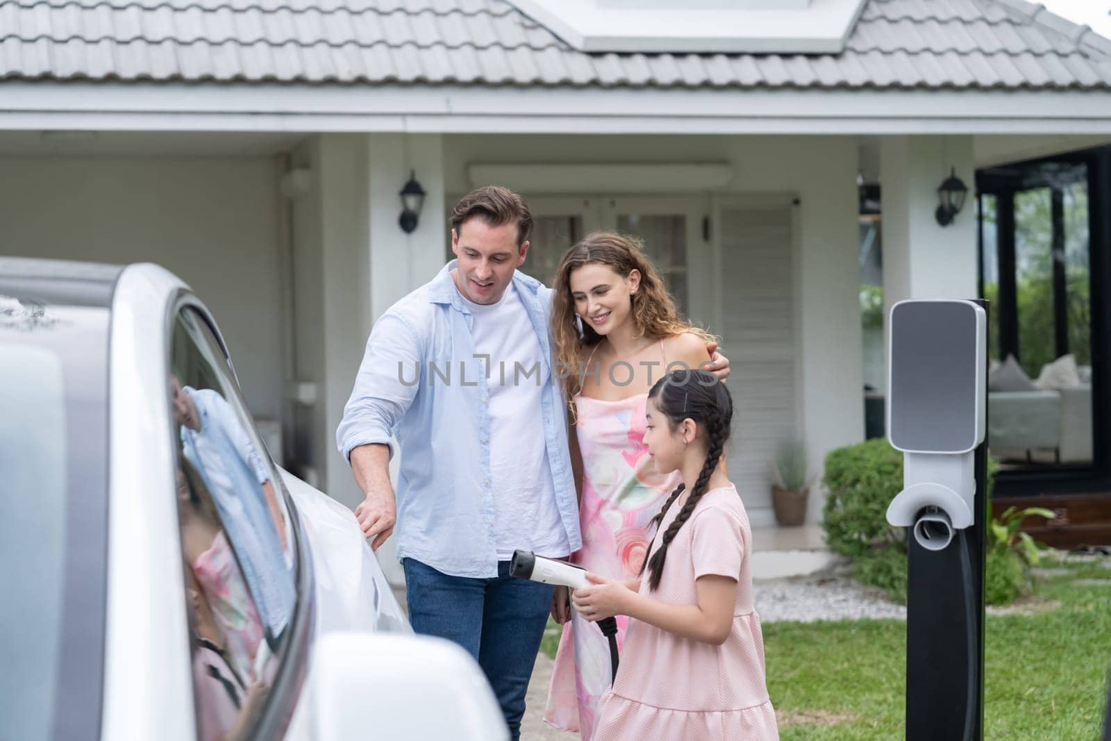 Happy little young girl learn about eco-friendly and energy sustainability as she help her family recharge electric vehicle from home EV charging station. EV car and modern family concept. Synchronos