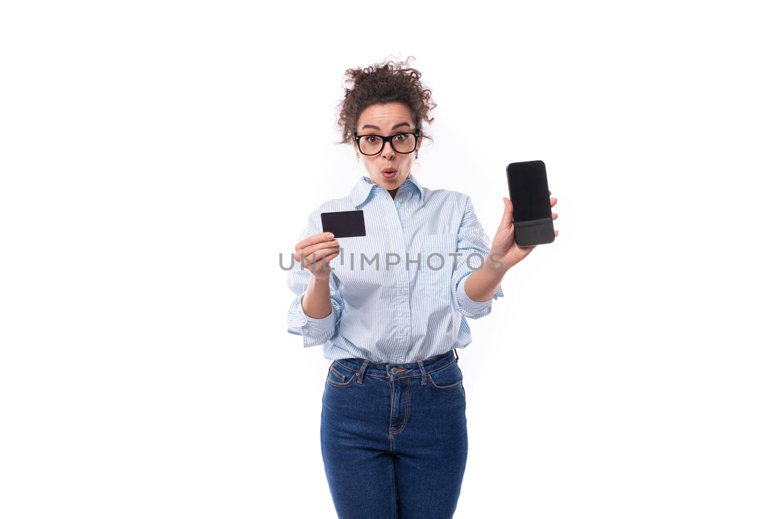 surprised young smiling stylish caucasian woman with curly hair holding plastic credit card and smartphone on white background with copy space.