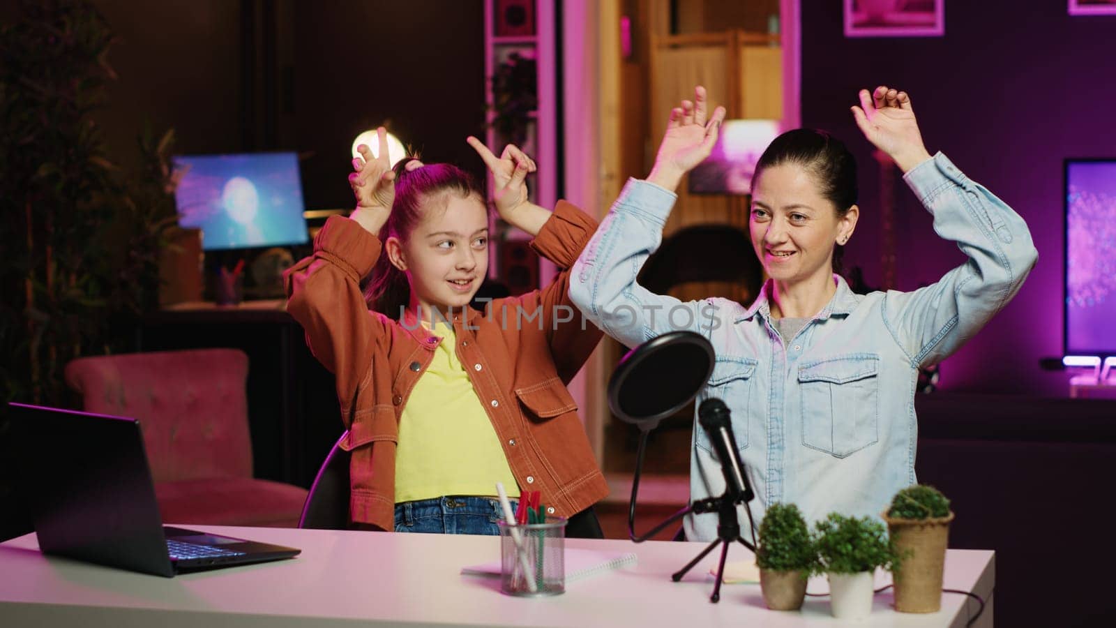 Cute kid teaching her mother how to do viral online dance moves while filming video for social media. Mom learning trending choreography from influencer daughter in apartment illuminated by pink neon