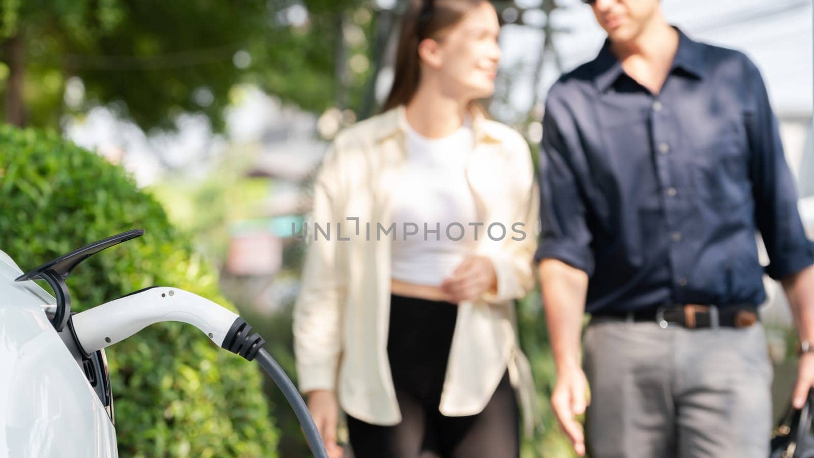 Young couple recharge electric car battery from charging station in green city park in springtime. Rechargeable EV car for sustainable environmental friendly urban travel lifestyle. Panorama Expedient