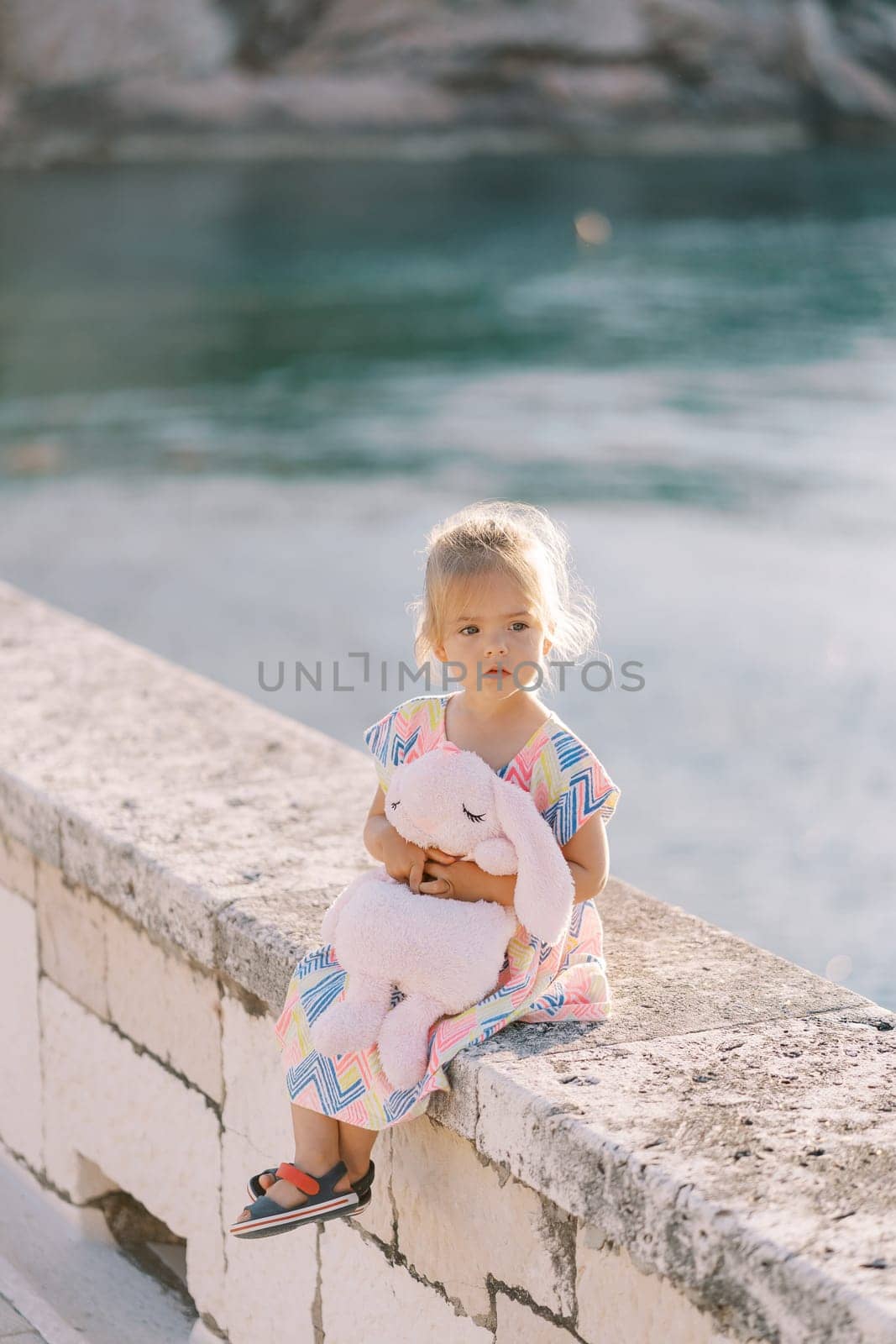 Little girl sits on a stone fence by the sea hugging a plush hare and looks away. High quality photo