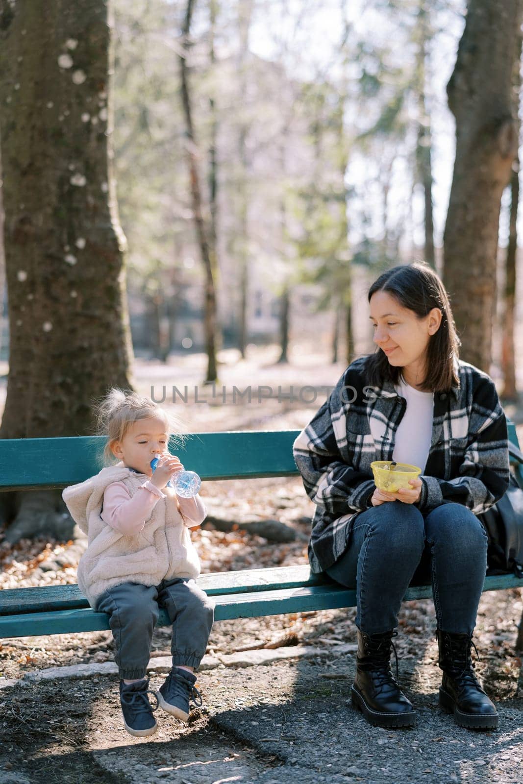 Little girl drinks water from a bottle while sitting next to her mother on a bench in a sunny park. High quality photo