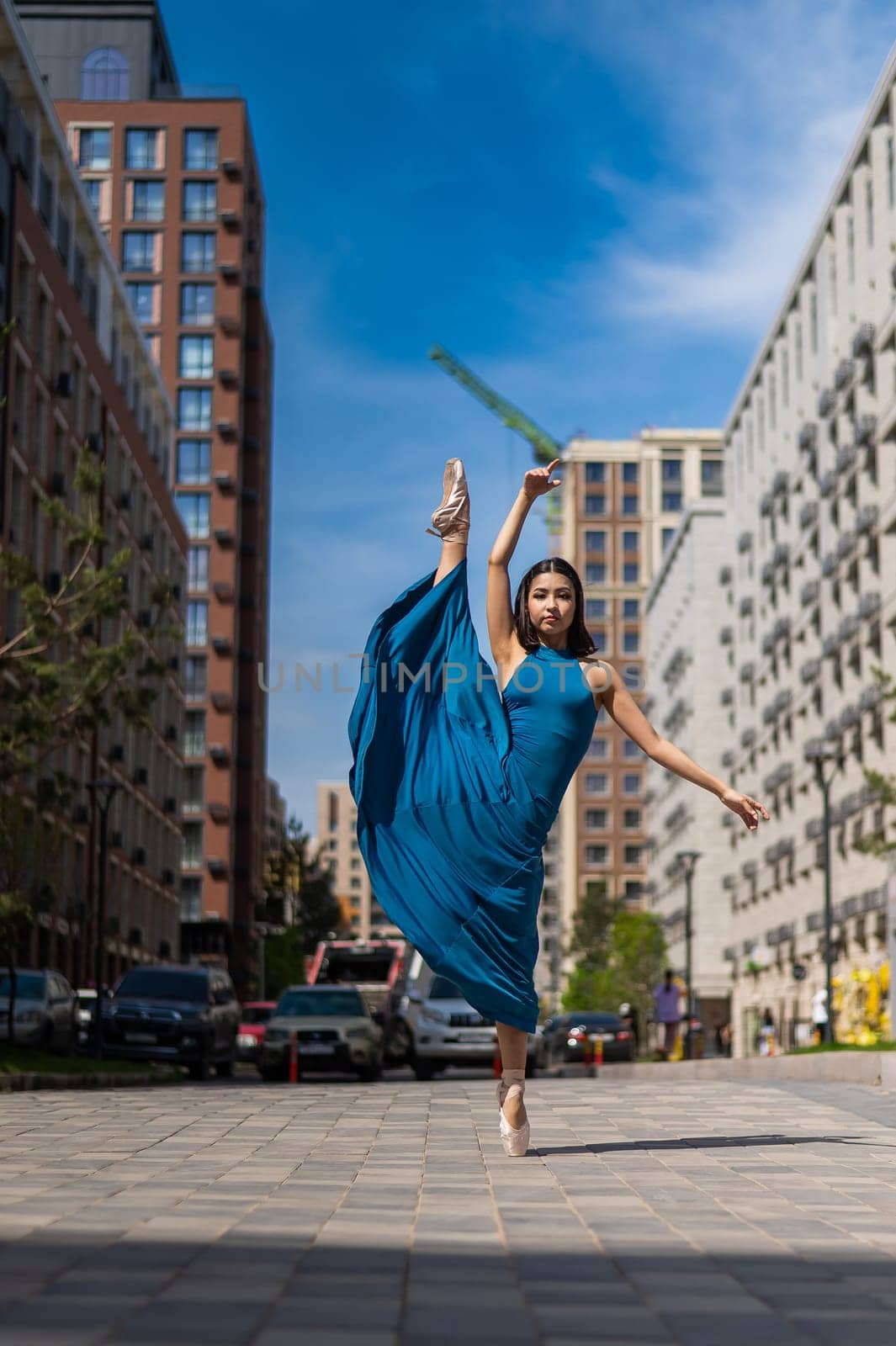 Beautiful Asian ballerina in blue dress posing in splits outdoors. Urban landscape. Vertical photo