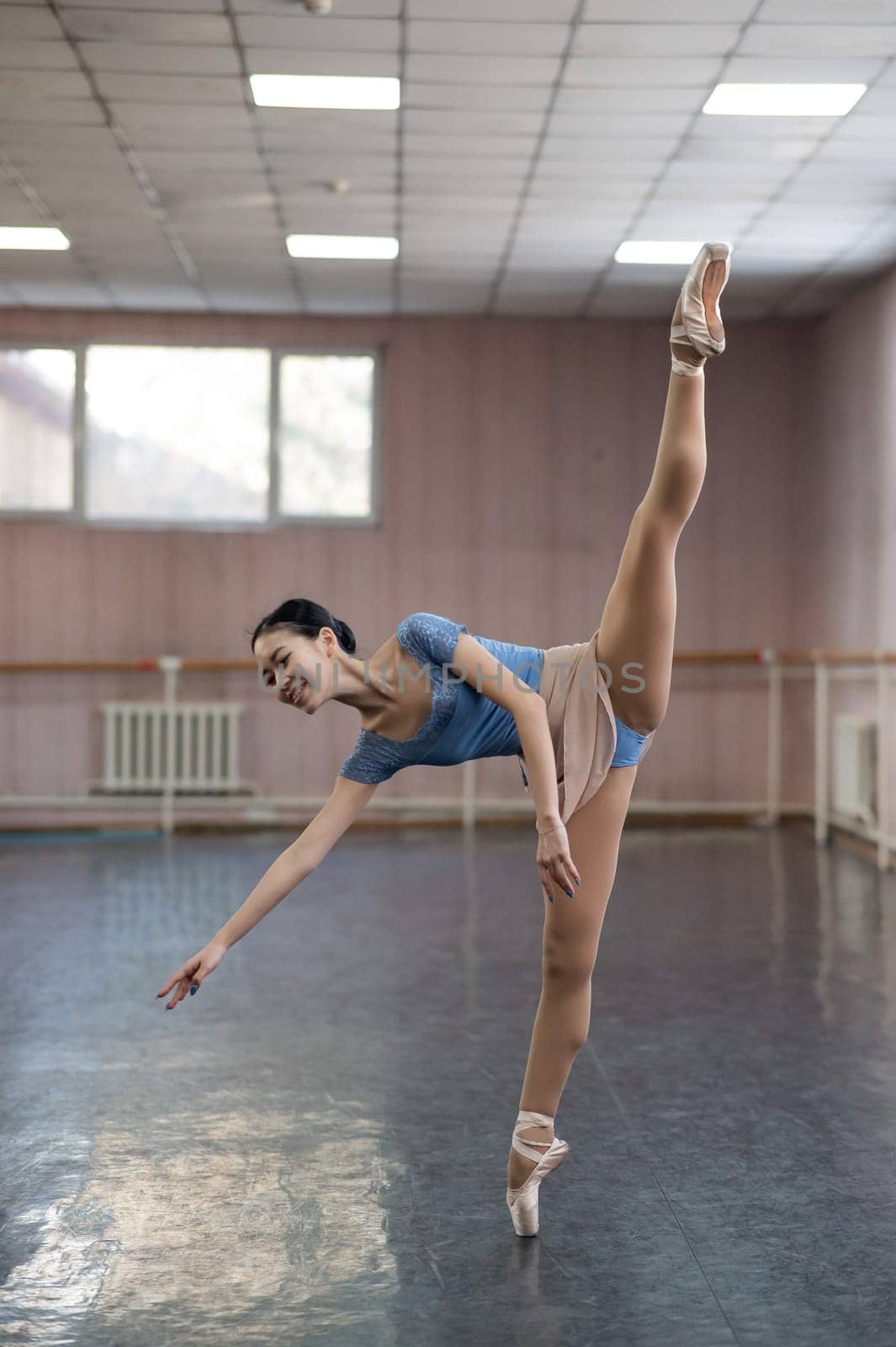 Graceful Asian ballerina in a blue bodysuit and a beige skirt is rehearsing in a dance class