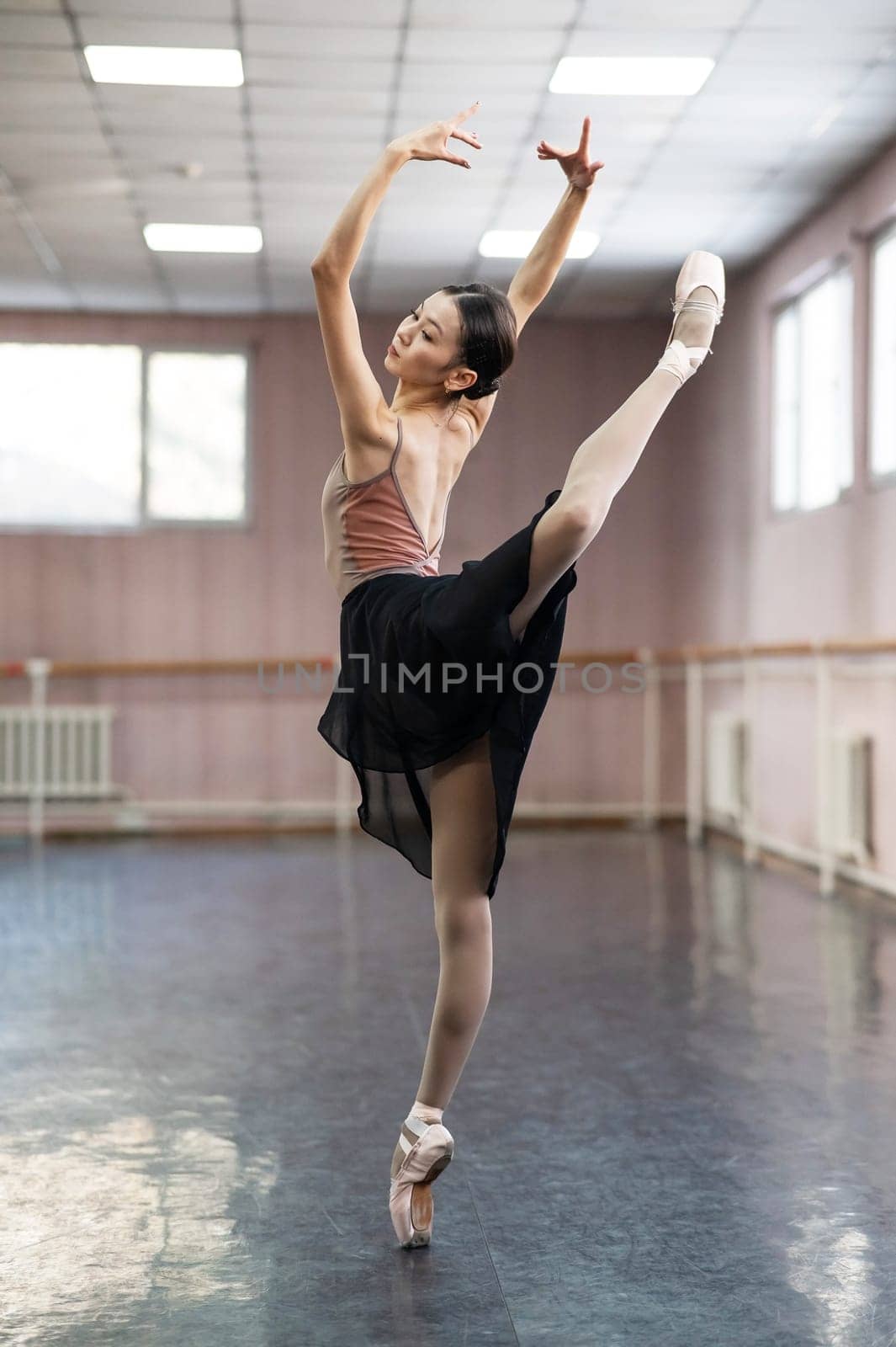 Graceful Asian ballerina in a beige bodysuit and black skirt is rehearsing in a dance class