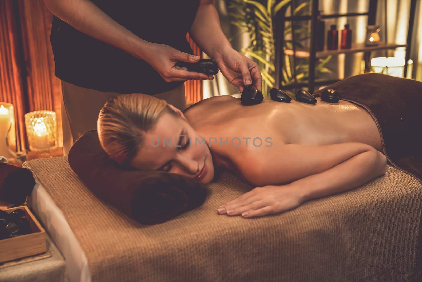 Hot stone massage at spa salon in luxury resort with warm candle light, blissful woman customer enjoying spa basalt stone massage glide over body with soothing warmth. Quiescent