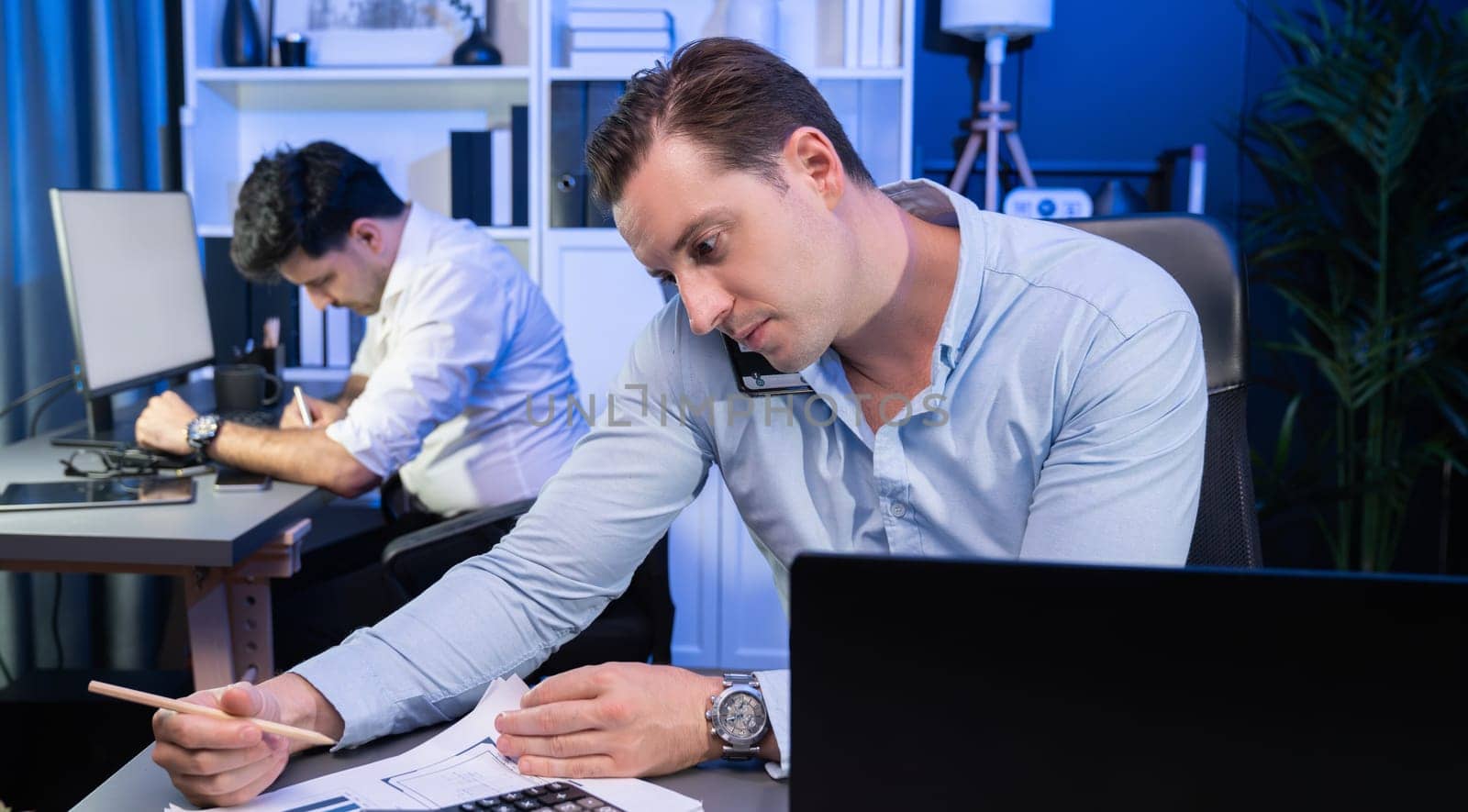 Serious businessman calling on phone with customer to create new project at night time, coworker writing document work sheet on desk. Concept of working on neon blue light modern office. Sellable.