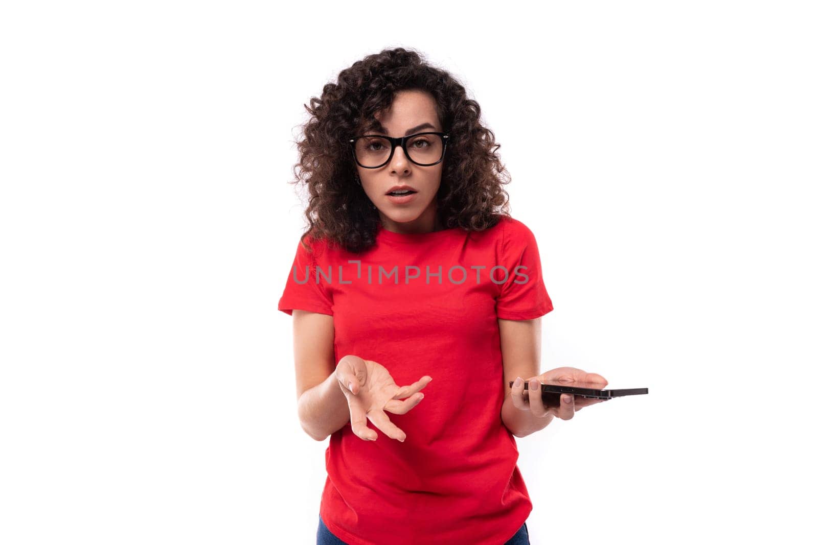caucasian young pretty curly brunette woman in a red t-shirt spreads her arms in excitement holding a smartphone in her hand by TRMK