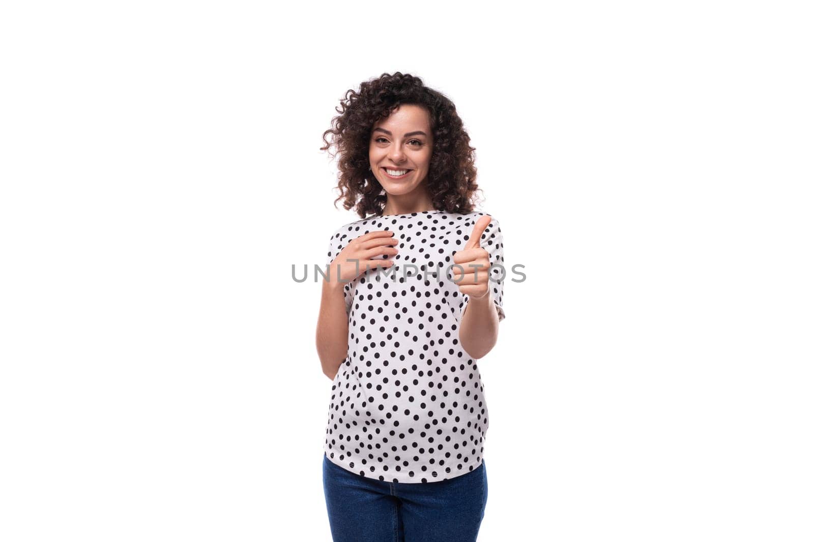 charming young brunette curly woman dressed in summer blouse with pea print points with index finger by TRMK