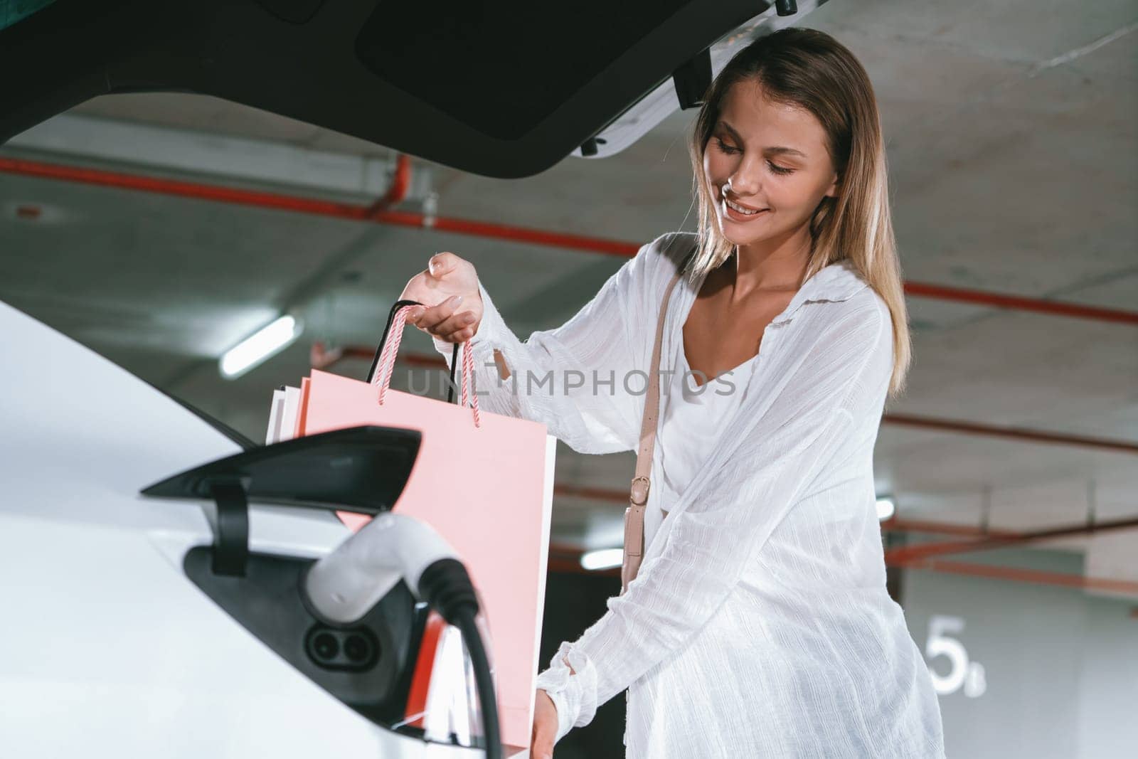 Young woman travel with EV electric car to shopping center parking lot charging in downtown city showing urban sustainability lifestyle by green clean rechargeable energy of electric vehicle innards