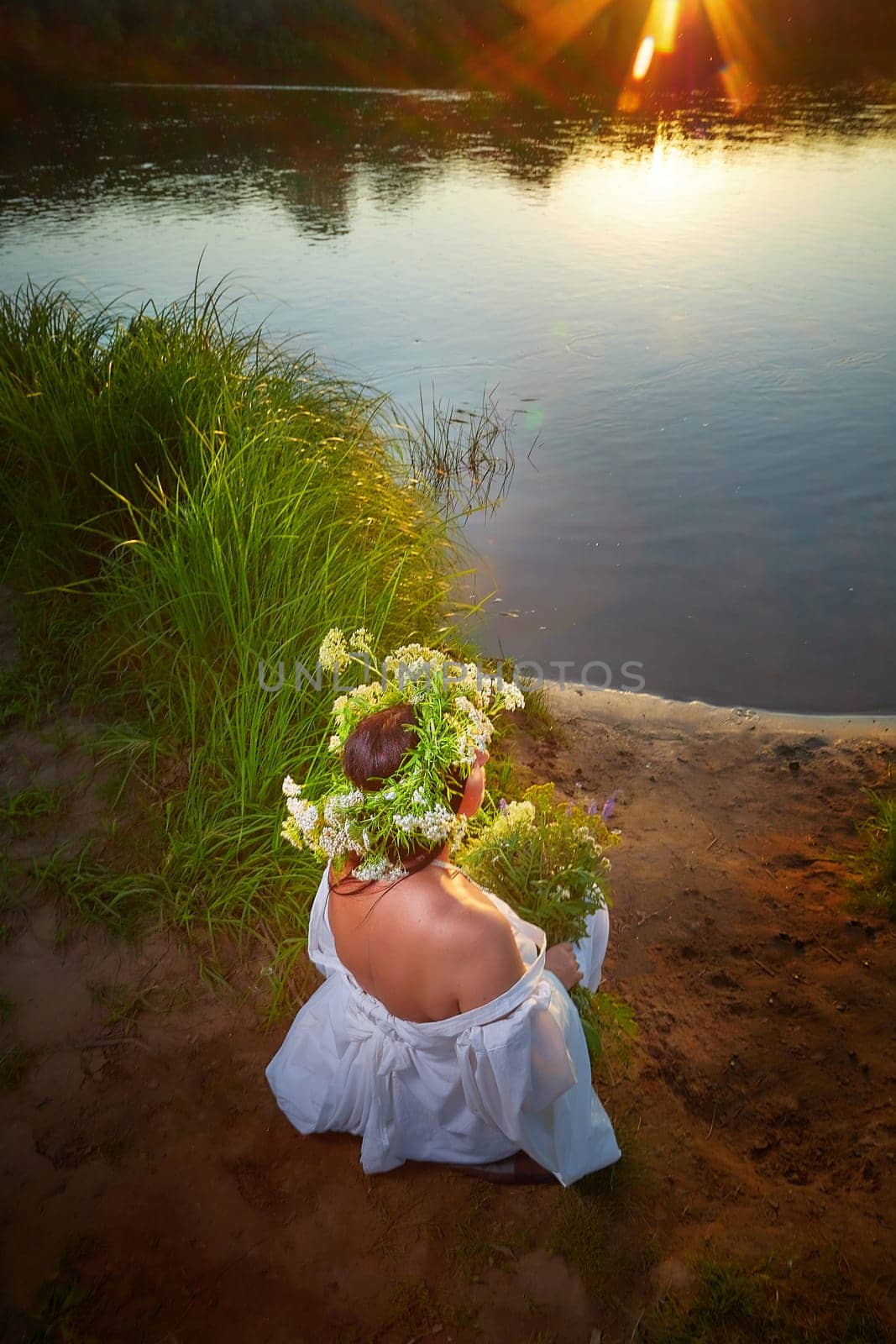 Adult mature brunette woman in a white dress, sundress and a wreath of flowers in summer by the water of river or lake in the evening at sunset. Celebration of the Slavic pagan holiday of Ivan Kupala by keleny