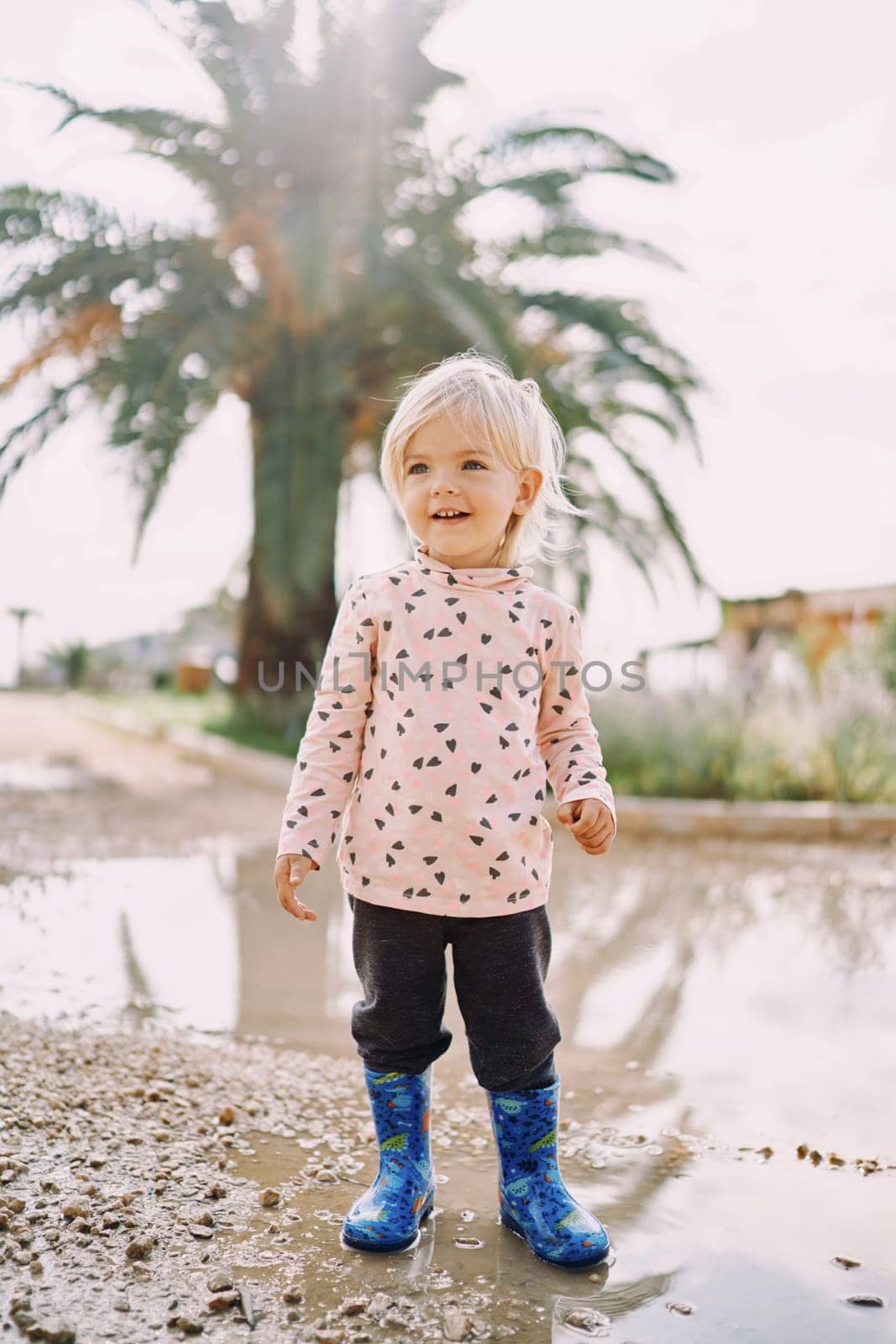 Smiling little girl in rubber boots stands in a puddle against the background of a green palm tree. High quality photo