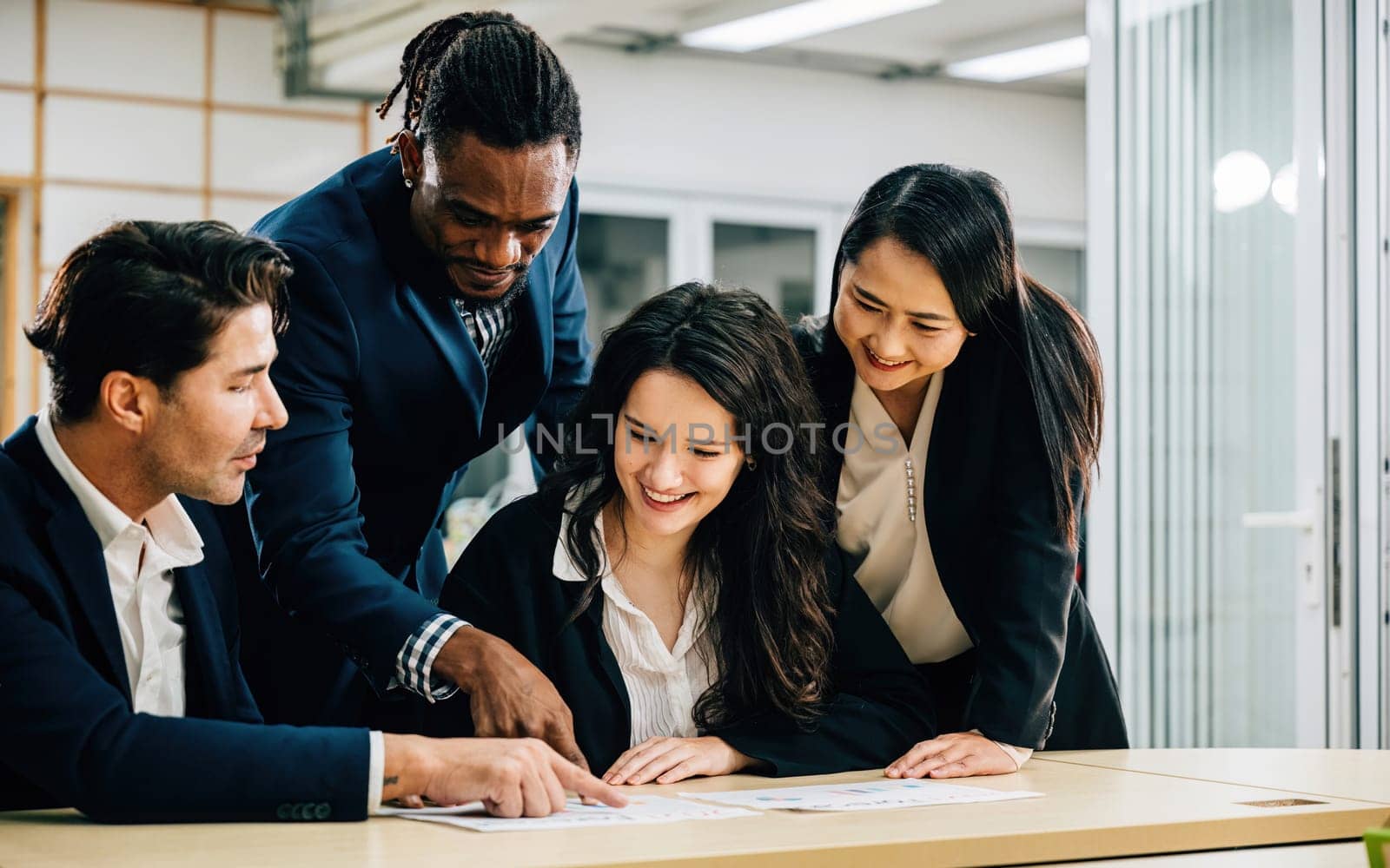 A diverse international team, with a female leader, collaborates in a meeting room. They work together, analyze graphs, and discuss strategies, displaying their commitment to success. Teamwork by Sorapop