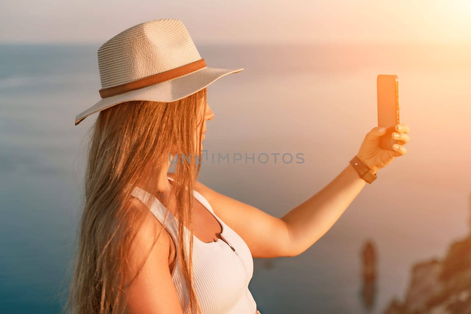 Selfie woman in a hat, white tank top, and shorts captures a selfie shot with her mobile phone against the backdrop of a serene beach and blue sea