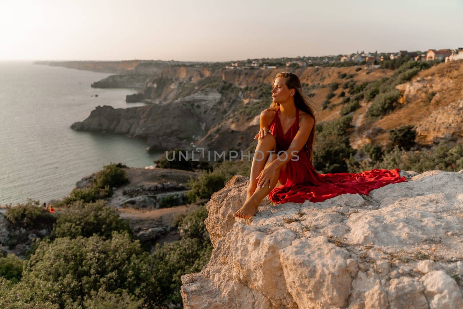 Woman sunset sea red dress, side view a happy beautiful sensual woman in a red long dress posing on a rock high above the sea on sunset