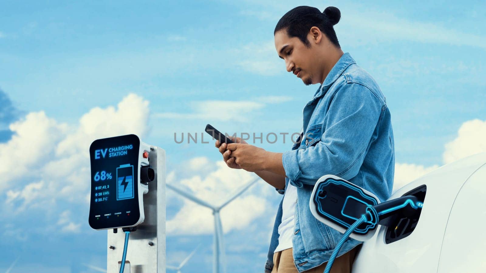 Asian man using smartphone while electric car recharging energy from EV charger from charging station at wind turbine farm. Alternative sustainable and clean energy utilization concept. Peruse