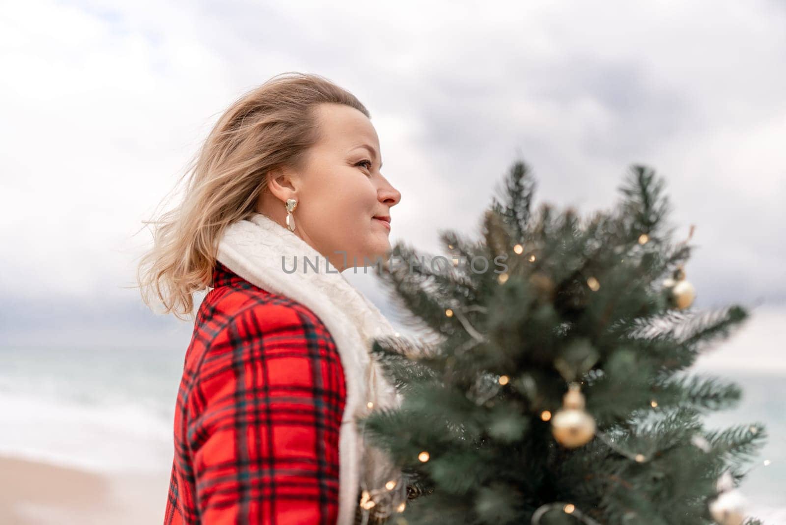 Blond woman holding Christmas tree by the sea. Christmas portrait of a happy woman walking along the beach and holding a Christmas tree in her hands. Dressed in a red coat, white suit. by Matiunina