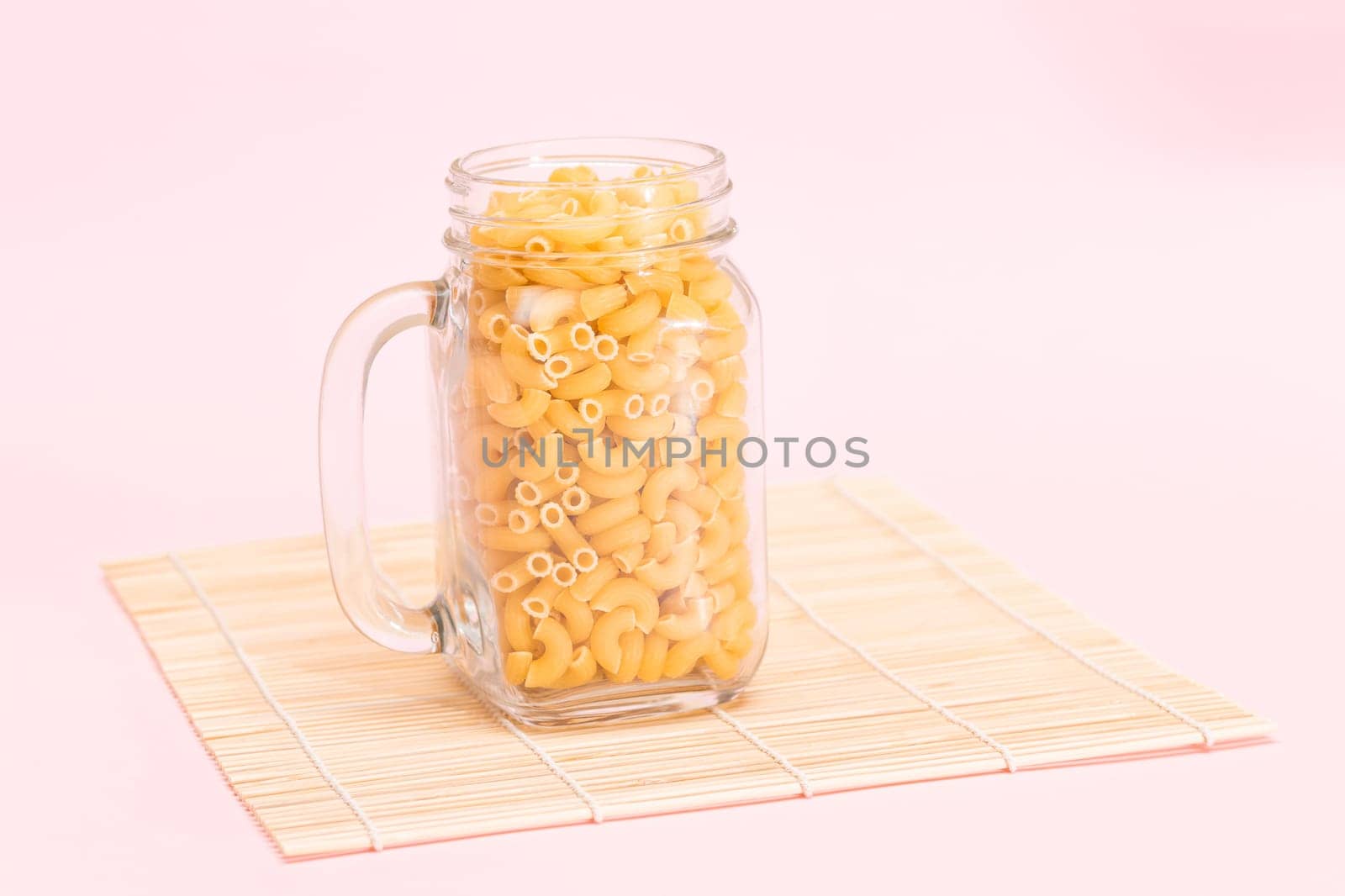 Uncooked Chifferi Rigati Pasta in Glass Jar on Bamboo Mat on Pink Background. Fat and Unhealthy Food. Scattered Classic Dry Macaroni. Italian Culture and Cuisine. Raw Pasta
