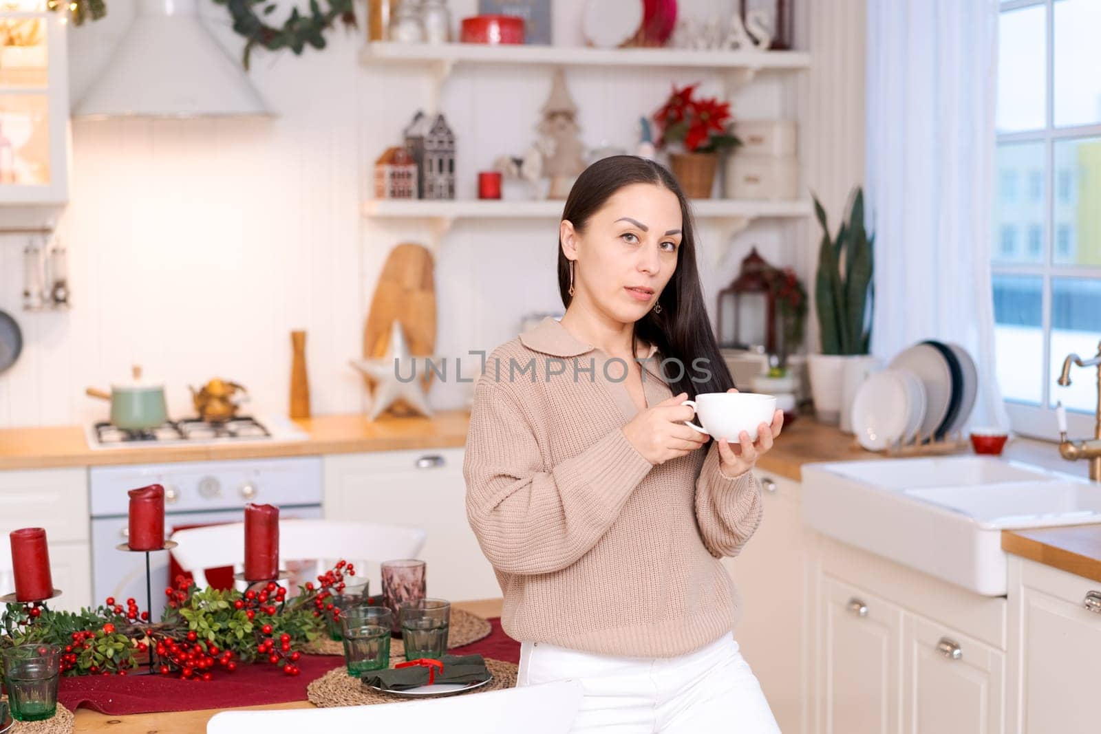 Concept festive Christmas atmosphere, cute woman drinking tea or coffee in a decorated kitchen wearing a sweater