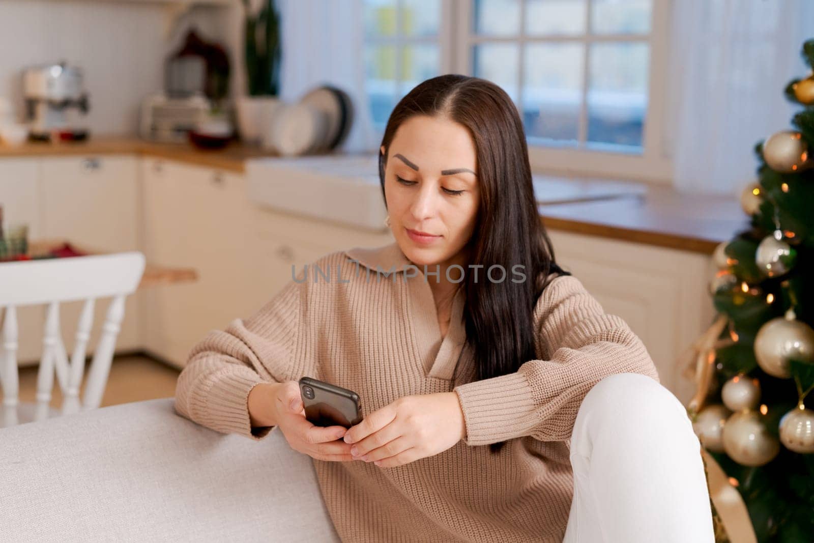 Woman with phone in hands sitting at home in kitchen on sofa during christmas by EkaterinaPereslavtseva