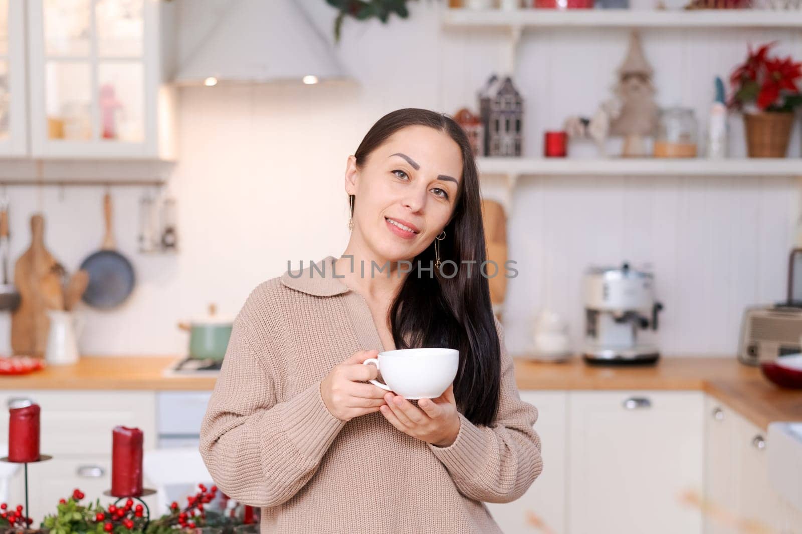 Concept festive Christmas atmosphere, cute woman drinking tea or coffee in a decorated kitchen wearing a sweater