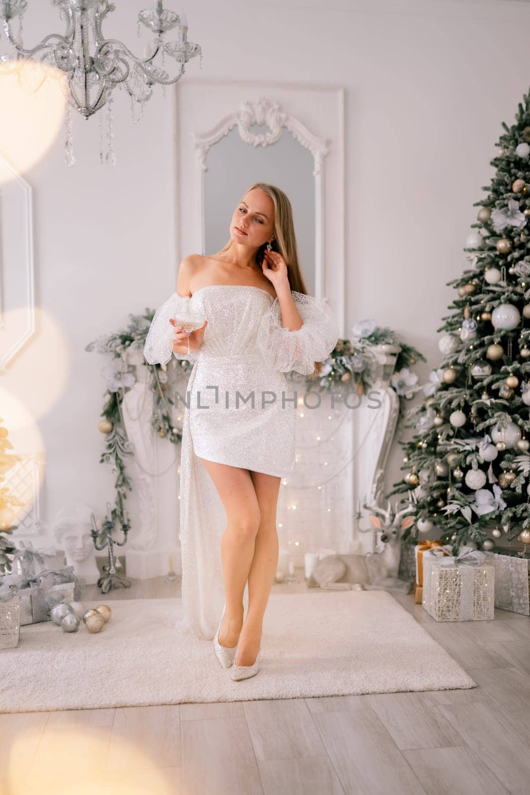 The blonde in the Christmas room. A beautiful blonde woman in a shiny light short dress with a train stands in a beautiful bright room decorated with a festive interior with a Christmas tree. by Matiunina
