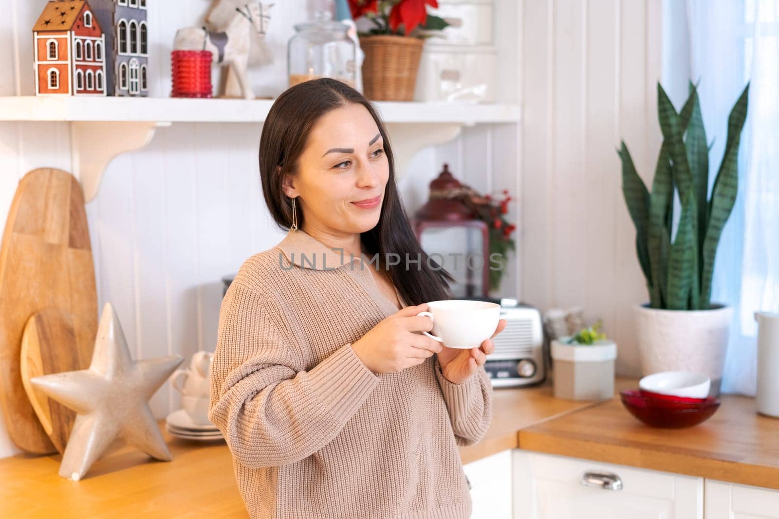 Concept festive Christmas atmosphere, cute woman drinking tea or coffee in a decorated kitchen wearing a sweater