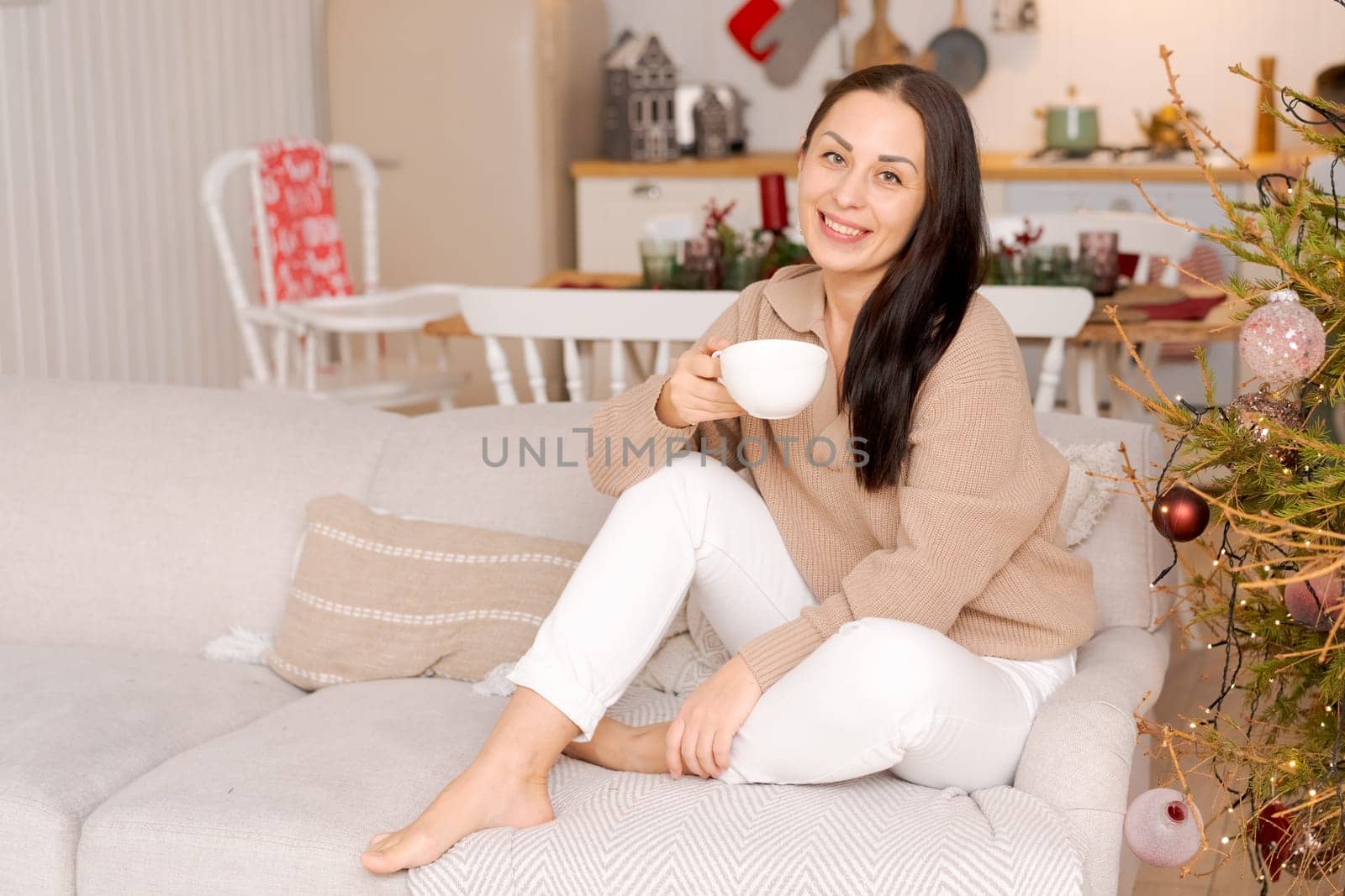 Concept festive Christmas atmosphere, lovely woman drinking tea or coffee sitting on a couch in a decorated kitchen wearing a sweater