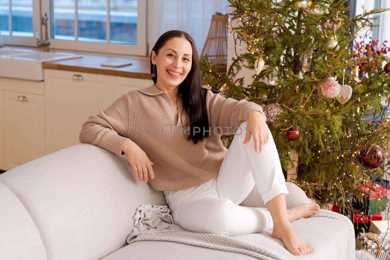 Happy woman sits on couch in sweater next to natural Christmas tree, holiday by EkaterinaPereslavtseva