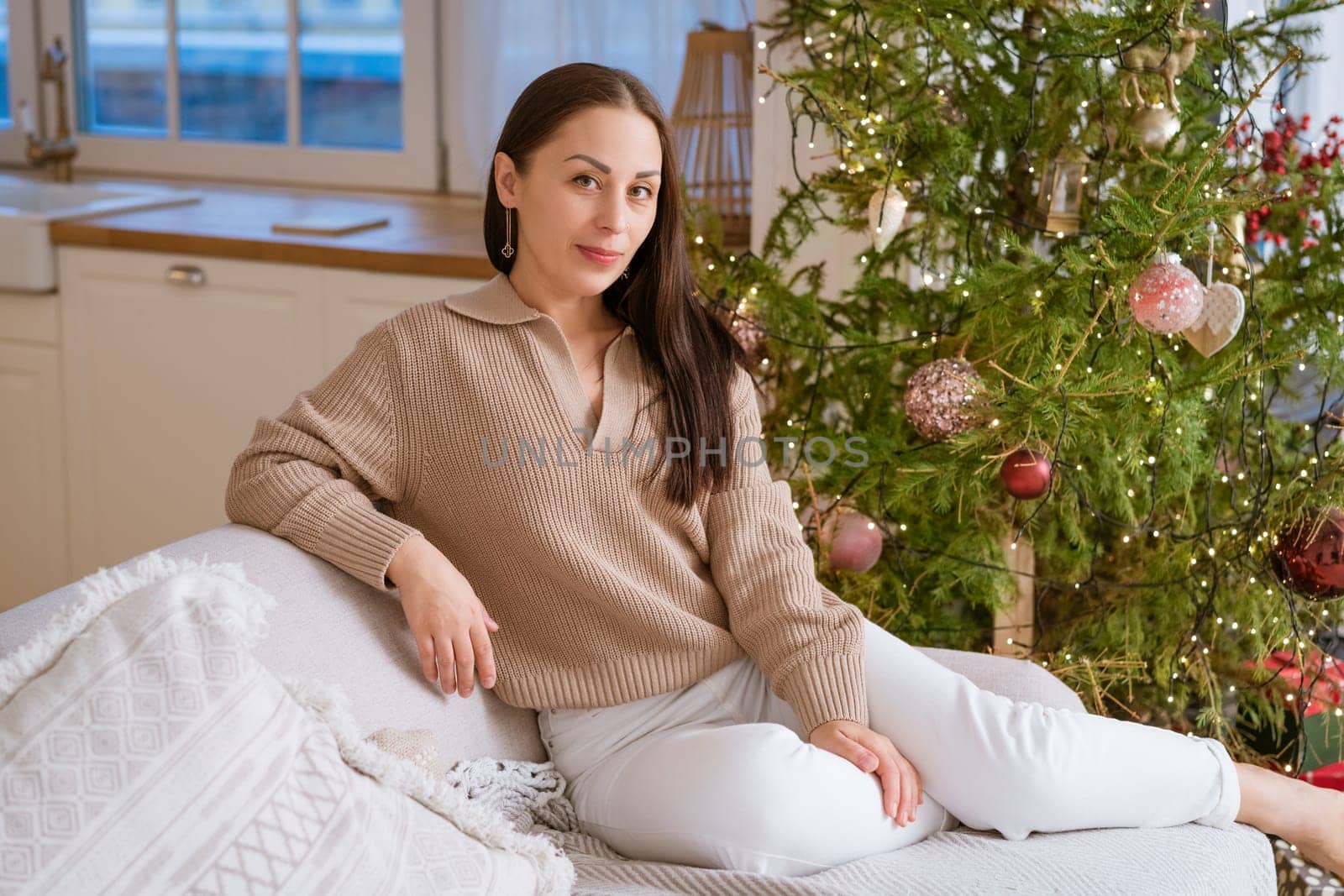 Happy woman sits on couch in sweater next to natural Christmas tree, holiday mood
