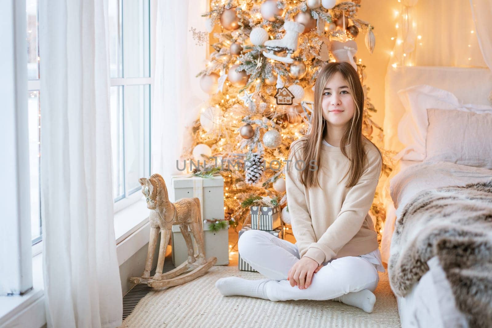 Cute girl sits on floor by window with Christmas tree in background in her bedroom