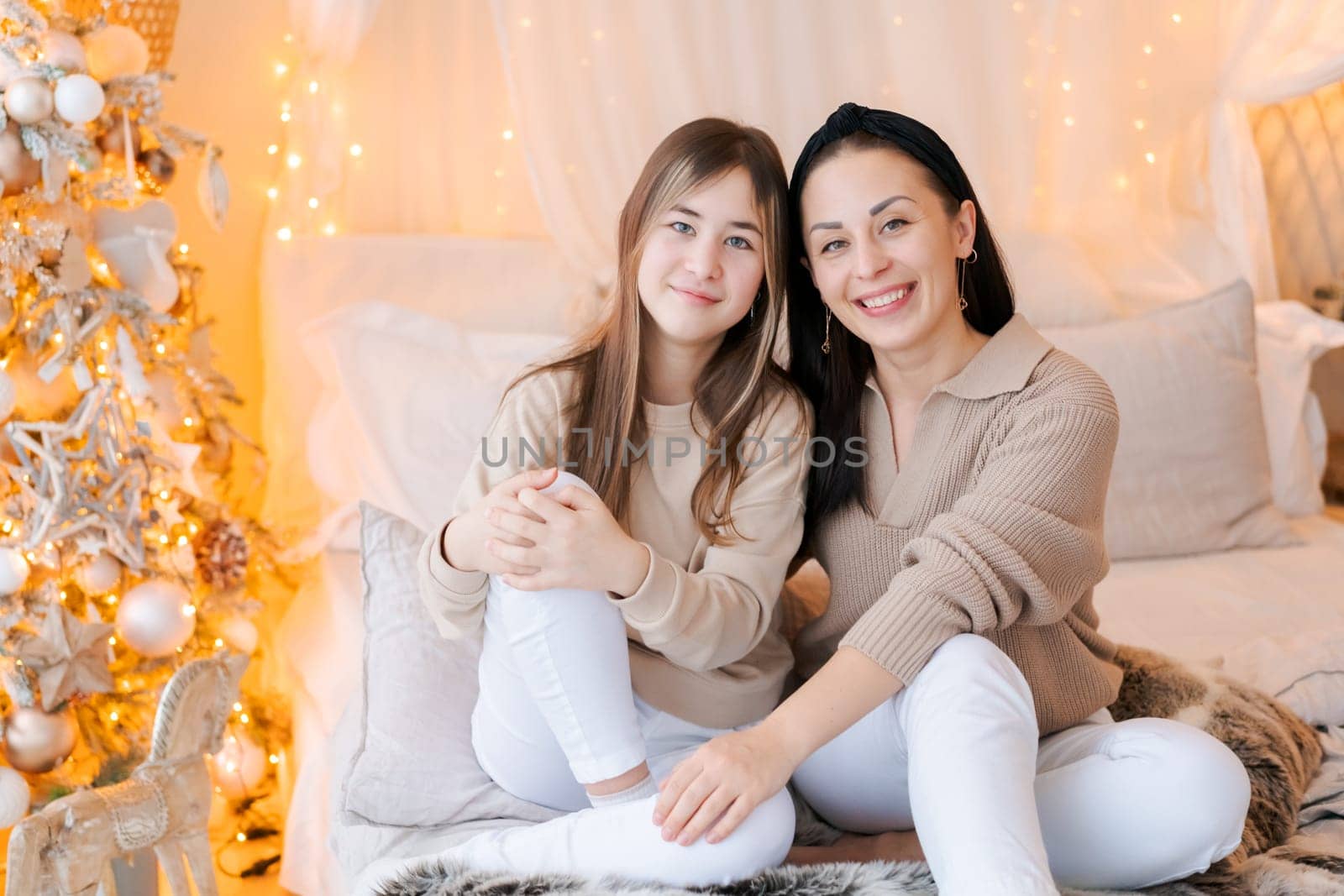 Happy mom and daughter cuddle on bed in decorated bedroom on Christmas Eve in anticipation of the party