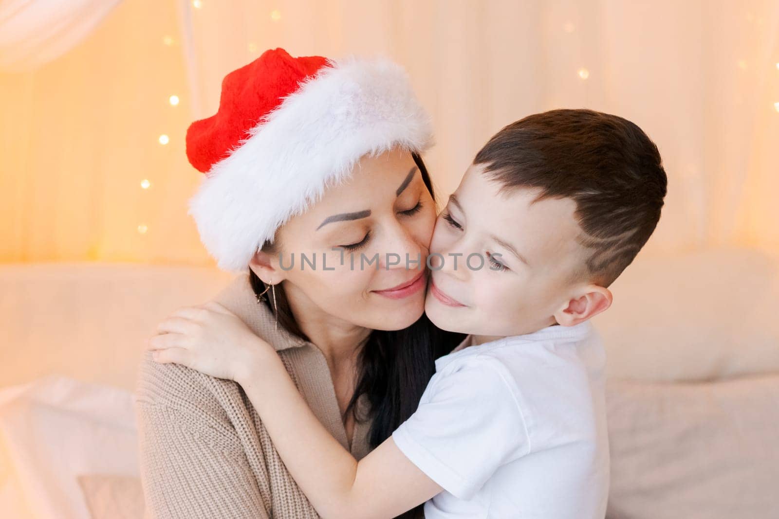 Happy mom and son wearing santa claus hat cuddle on bed in decorated bedroom by EkaterinaPereslavtseva